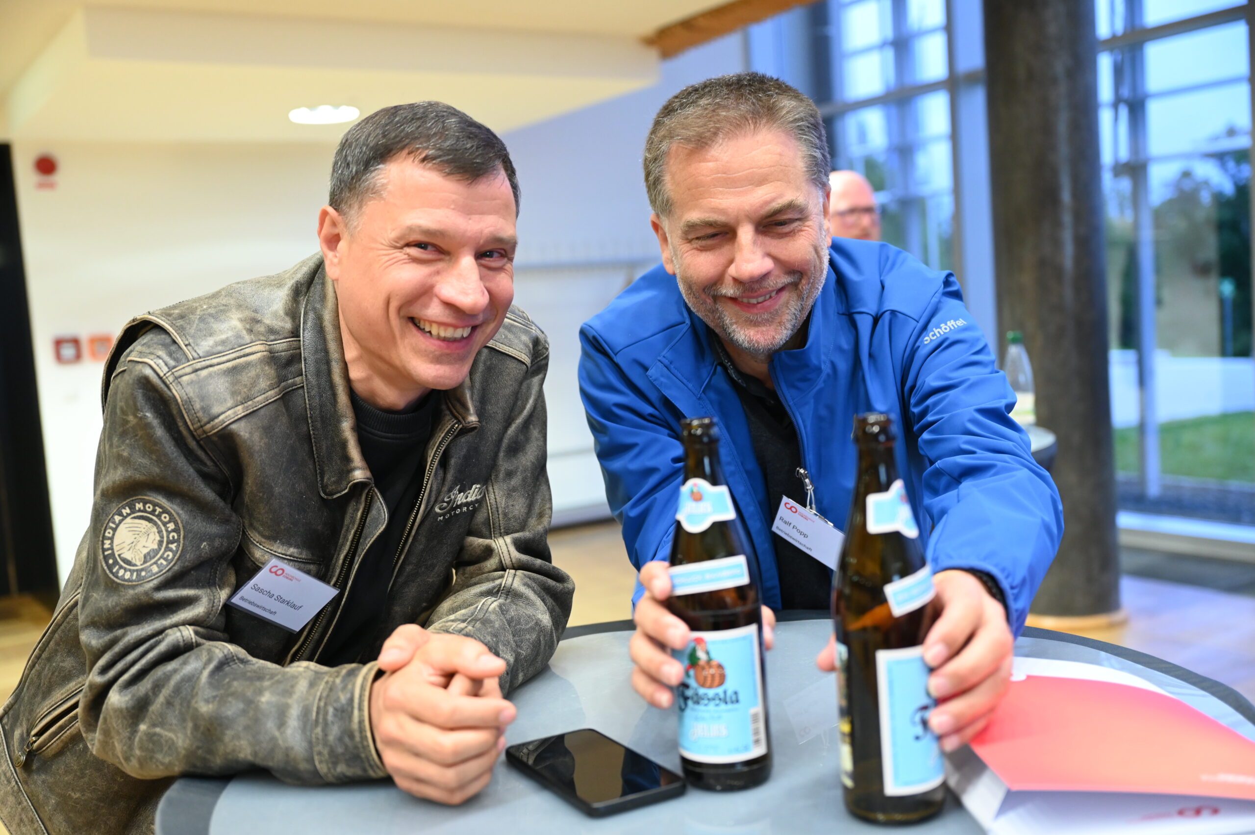 Zwei lächelnde Männer, die sich über einen runden Tisch in einer Hochschule beugen, jeder hält eine Bierflasche. Auf dem Tisch liegen ein Smartphone und ein roter Ordner. Im Hintergrund sind große Fenster zu sehen, die den lebendigen Geist einer Silberjubiläumsfeier einfangen. at Hochschule Coburg