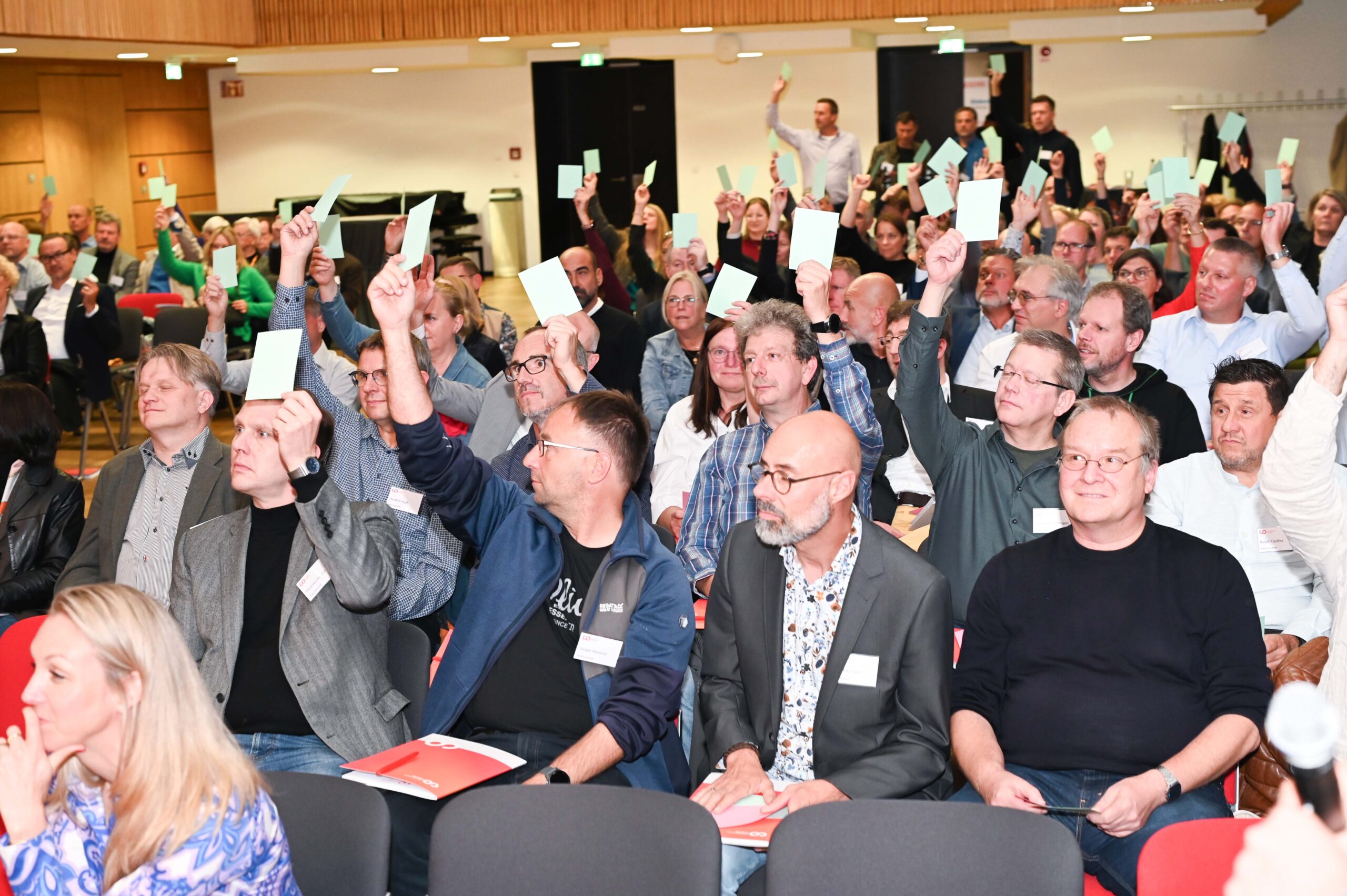 Eine große Gruppe von Menschen, die in einem Hörsaal sitzt, hebt grüne Karten und nimmt an einer Abstimmung oder einem Entscheidungsprozess im Silberjubiläum der Hochschule teil. Die meisten Gesichter sehen aufmerksam und engagiert aus, und viele Hände halten die Karten in die Höhe. at Hochschule Coburg