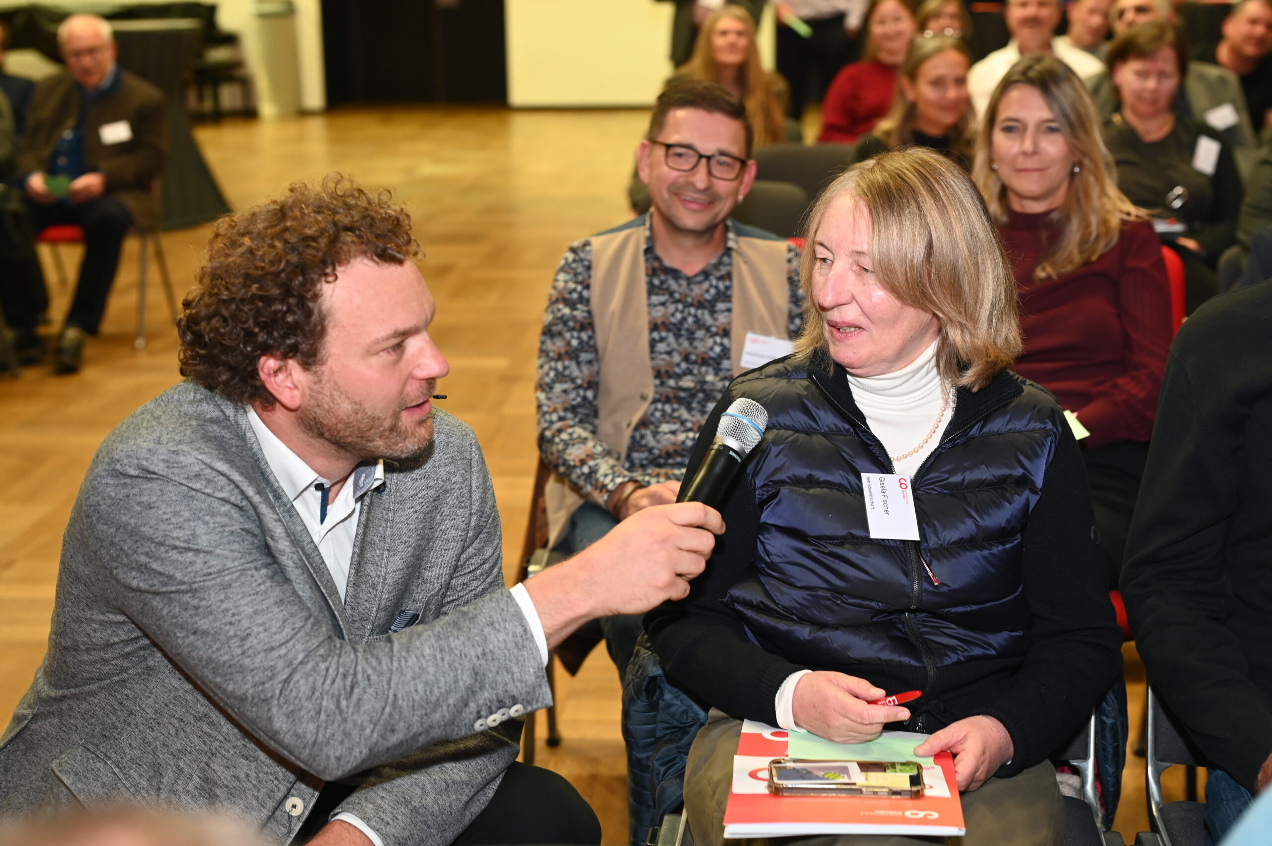 Bei einem Silberjubiläum der Hochschule hält ein Mann in einem grauen Blazer ein Mikrofon in Richtung einer blonden Frau im Publikum, während andere in der lebhaften Halle lächelnd zusehen. at Hochschule Coburg
