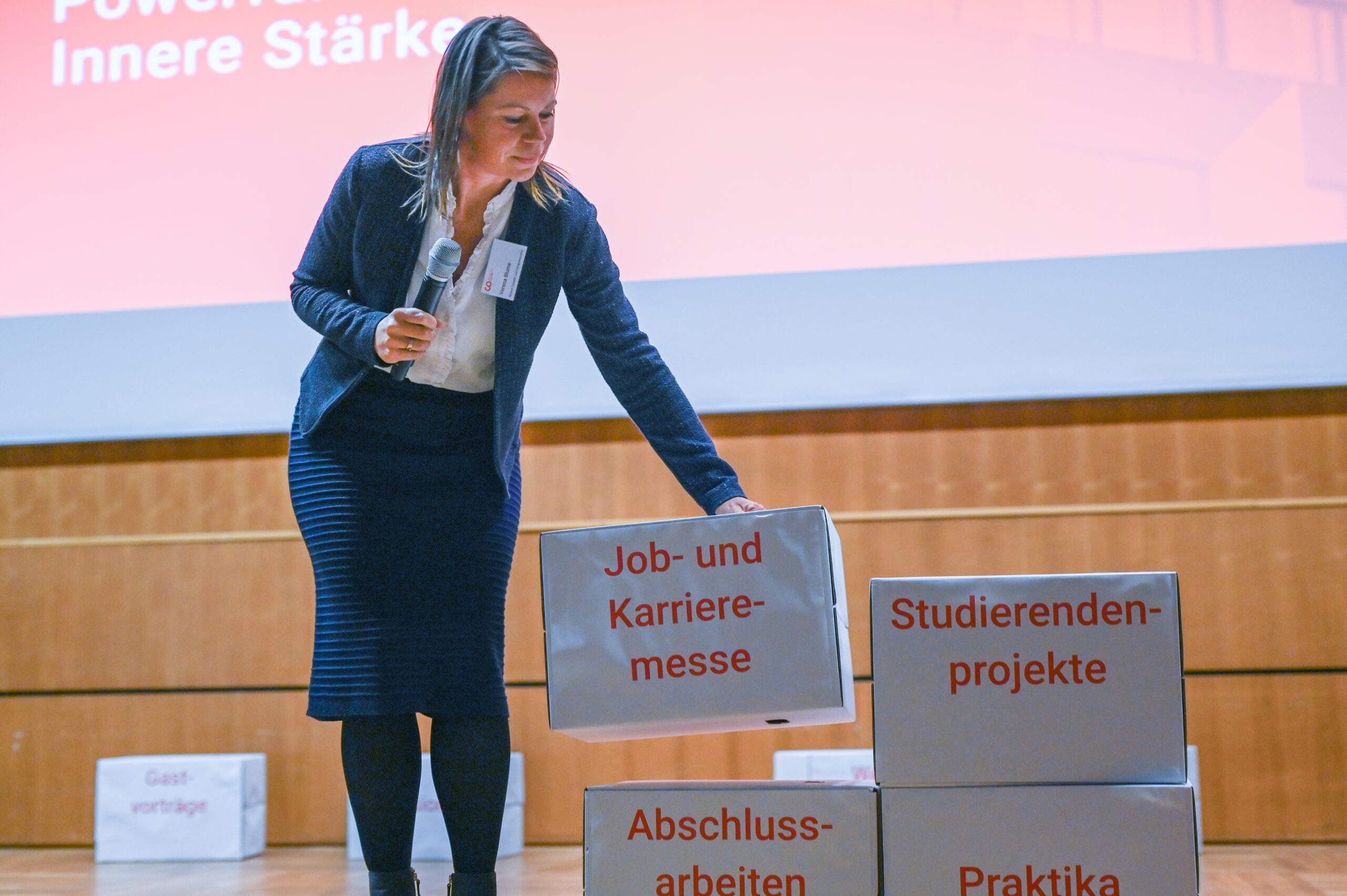 Eine Frau in Business-Kleidung hält ein Mikrofon in der Hand und ordnet große Kisten mit deutschen Begriffen wie "Job- und Karrieremesse" und "Studierendenprojekte" auf der Bühne der Hochschule an, während im Hintergrund eine Präsentationswand zu sehen ist. at Hochschule Coburg