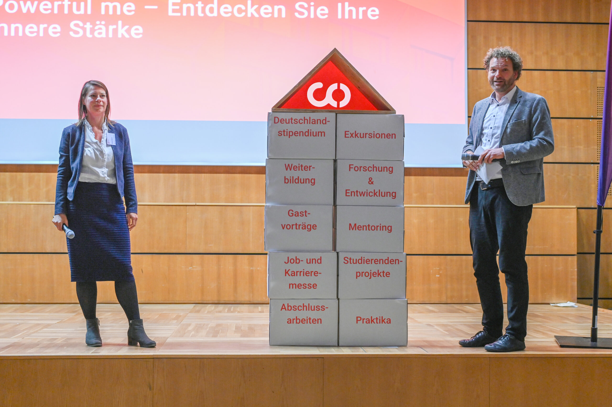 Zwei Personen stehen auf einer Bühne neben einer großen Blockstruktur mit deutschen Worten über Bildung und Karriere, die mit einem roten Pfeil und einem Unendlichkeitssymbol versehen sind und das Silberjubiläum der Hochschule hervorheben. Im Hintergrund ist ein Präsentationsbildschirm zu sehen. at Hochschule Coburg