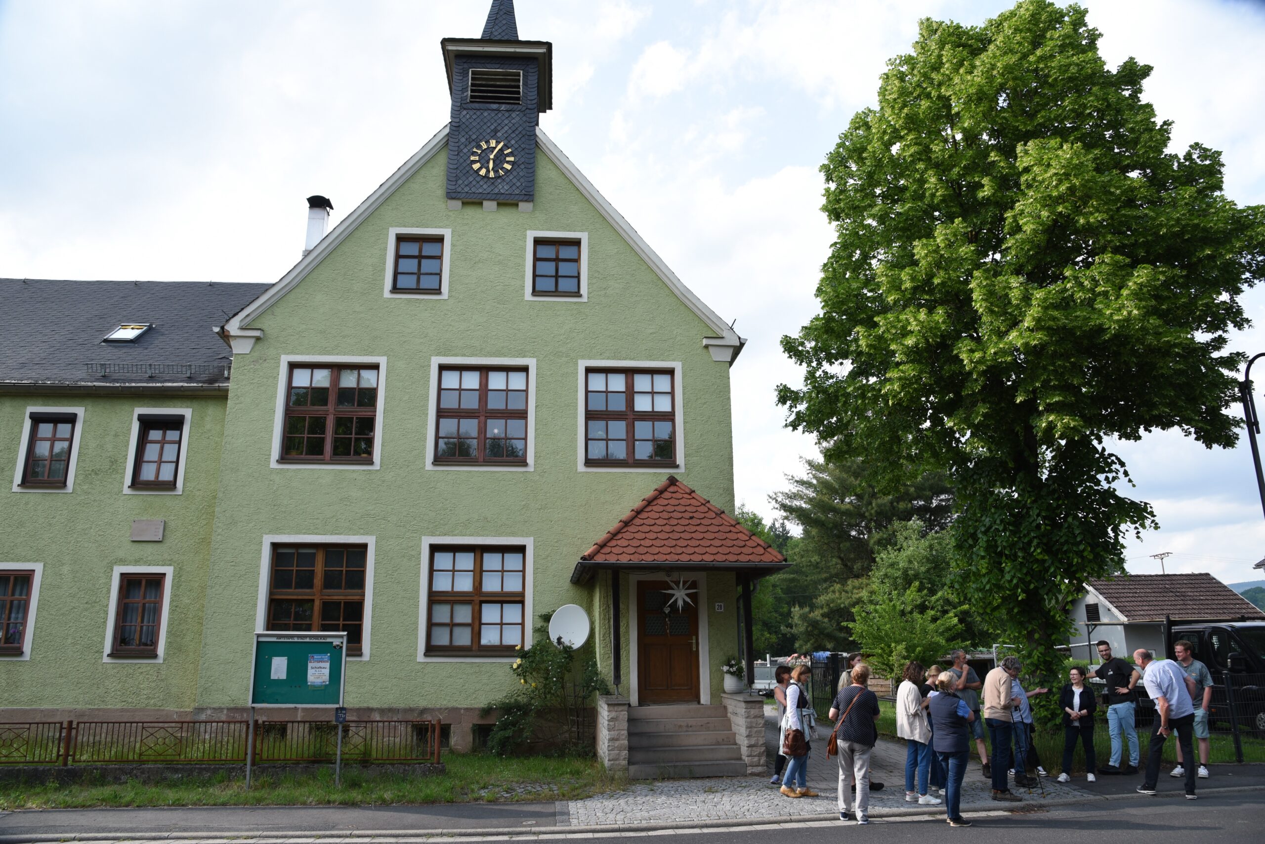 Eine Gruppe von Menschen steht und unterhält sich auf einem kopfsteingepflasterten Weg vor einem grünen Gebäude mit großen Fenstern, einem Uhrenturm und einem Baum in der Nähe. Die ruhige, gesellige Szene wirkt wie ein automatisch gespeicherter Entwurf des täglichen Lebens. at Hochschule Coburg