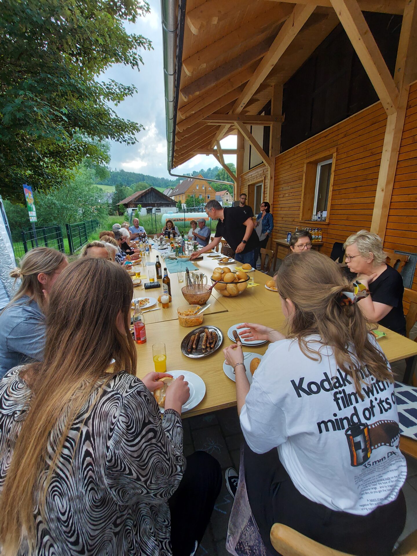 Eine Gruppe von Menschen versammelt sich um einen langen Tisch im Freien und teilt eine Mahlzeit mit Brot, Würstchen und Getränken. In der Nähe eines Holzhauses und üppigen Grüns liegt Lachen in der Luft - ein automatisch gespeicherter Entwurf fröhlicher Momente unter einem bewölkten Himmel. at Hochschule Coburg