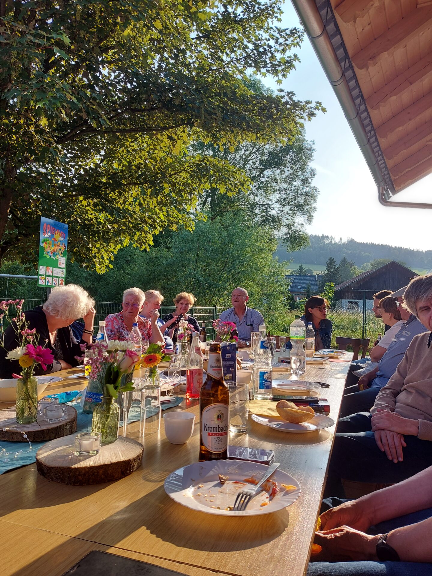 Eine Gruppe von Menschen sitzt um einen langen Holztisch im Freien mit Essen, Getränken und Blumen und genießt einen sonnigen Tag in einem Garten mit Bäumen und Hügeln - die Essenz eines automatisch gespeicherten Entwurfs von Sommererinnerungen. at Hochschule Coburg