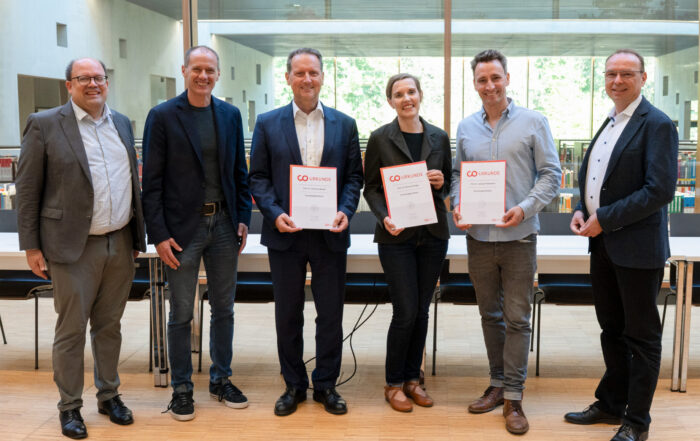 Sechs Erwachsene stehen in einem Raum vor einem Tisch, drei von ihnen halten Urkunden oder Auszeichnungen in der Hand, alle lächeln in die Kamera und freuen sich über neue Professuren. Im Hintergrund sind Bücherregale und große Fenster mit Grünpflanzen zu sehen. at Hochschule Coburg