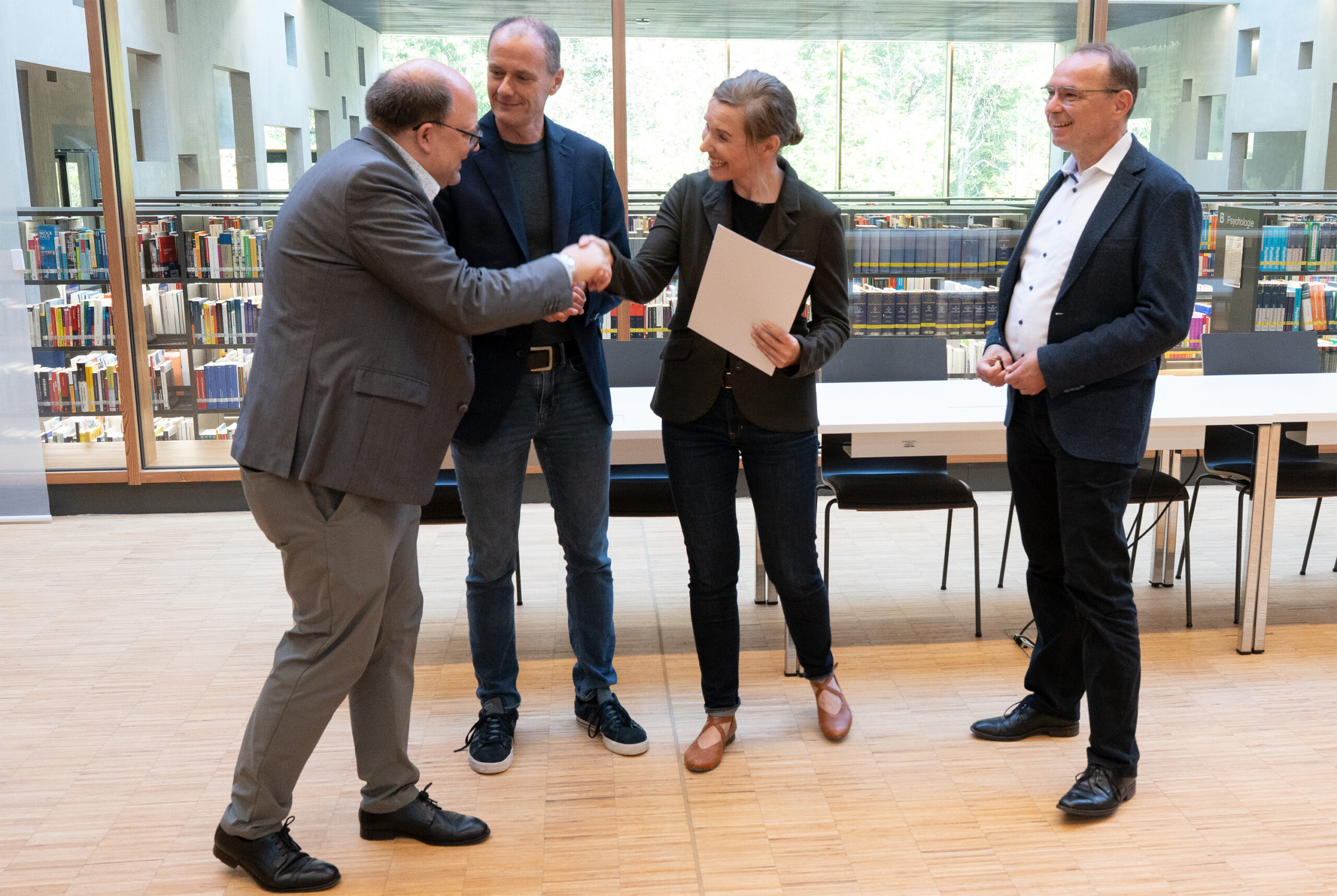 Vier Personen stehen in einer hellen Bibliothek zusammen. Ein Mann im Anzug schüttelt einer lächelnden Frau die Hand, die ein Dokument mit der Aufschrift "Forschungsprofessuren" in der Hand hält, während zwei weitere Personen beobachten und im Sinne von "Wissen für die Zukunft" teilen. at Hochschule Coburg