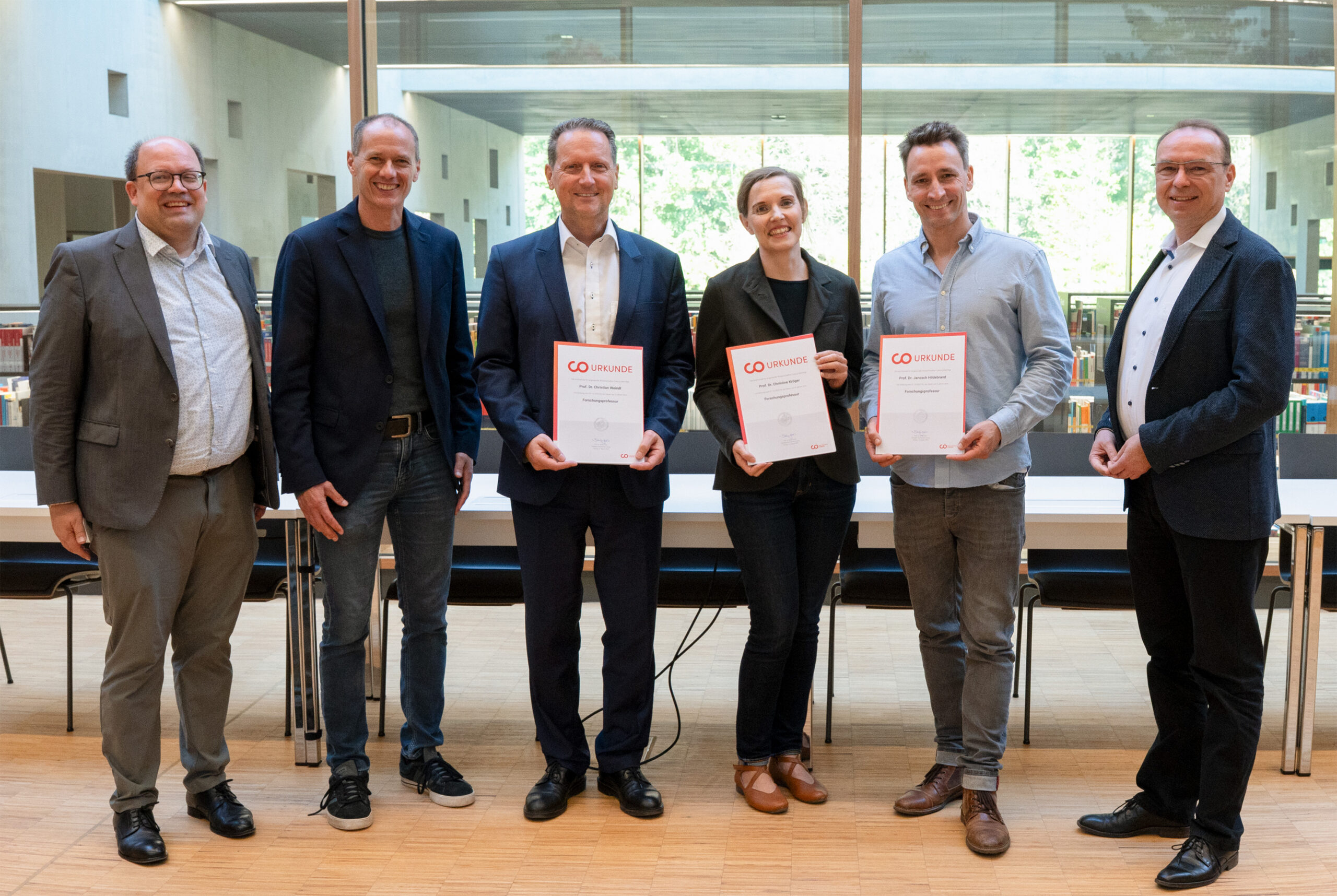 Sechs Erwachsene stehen in einem Raum vor einem Tisch, drei von ihnen halten Urkunden oder Auszeichnungen in der Hand, alle lächeln in die Kamera und freuen sich über neue Professuren. Im Hintergrund sind Bücherregale und große Fenster mit Grünpflanzen zu sehen. at Hochschule Coburg