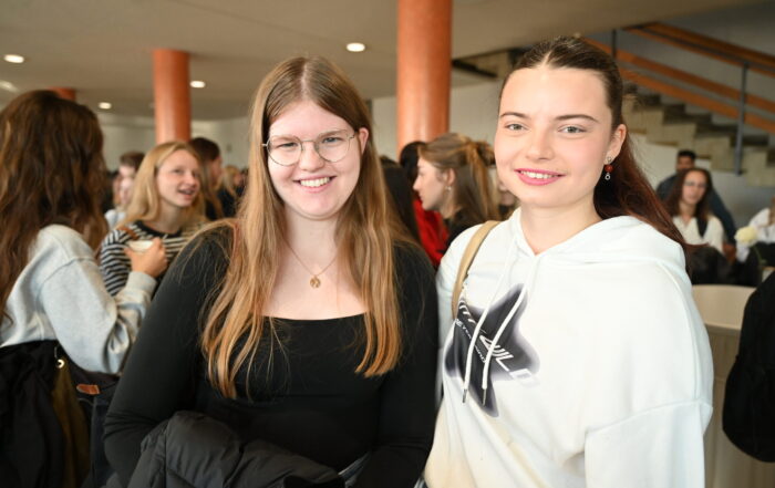 Zwei junge Frauen stehen lächelnd zusammen in einer belebten Halle in Coburg, Bayern - bekannt als die günstigste Hochschulstadt. Die eine Frau hat langes glattes Haar und eine Brille, die andere hat ihr Haar zurückgebunden und trägt einen weißen Kapuzenpullover. at Hochschule Coburg