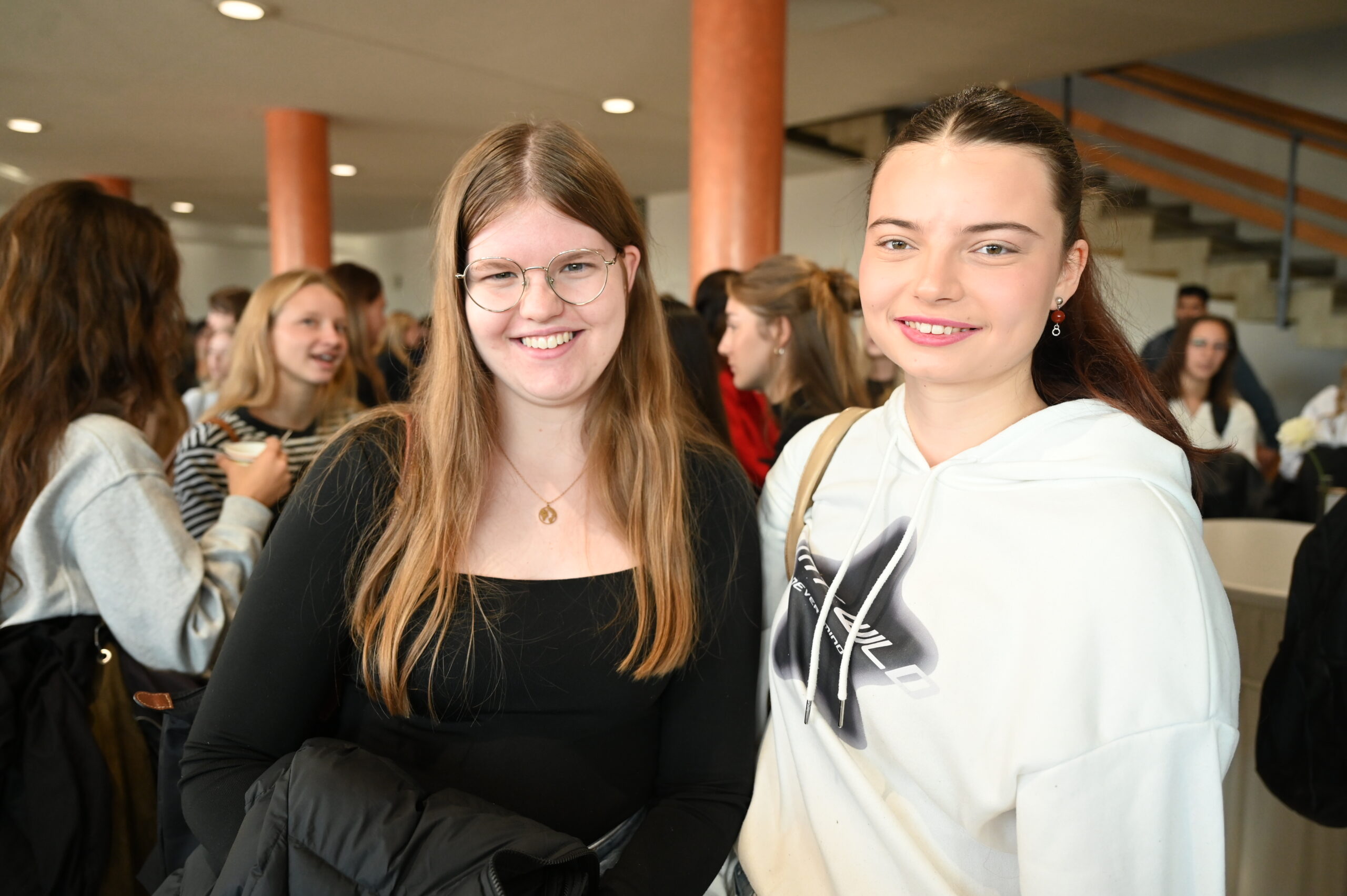 Zwei junge Frauen stehen lächelnd zusammen in einer belebten Halle in Coburg, Bayern - bekannt als die günstigste Hochschulstadt. Die eine Frau hat langes glattes Haar und eine Brille, die andere hat ihr Haar zurückgebunden und trägt einen weißen Kapuzenpullover. at Hochschule Coburg
