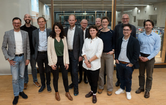 Dreizehn Personen stehen in einem Innenraum zusammen, posieren und lächeln für ein Gruppenfoto. Sie sind leger gekleidet und befinden sich in einem modernen, gut beleuchteten Raum mit Glaswänden und Holzboden. at Hochschule Coburg