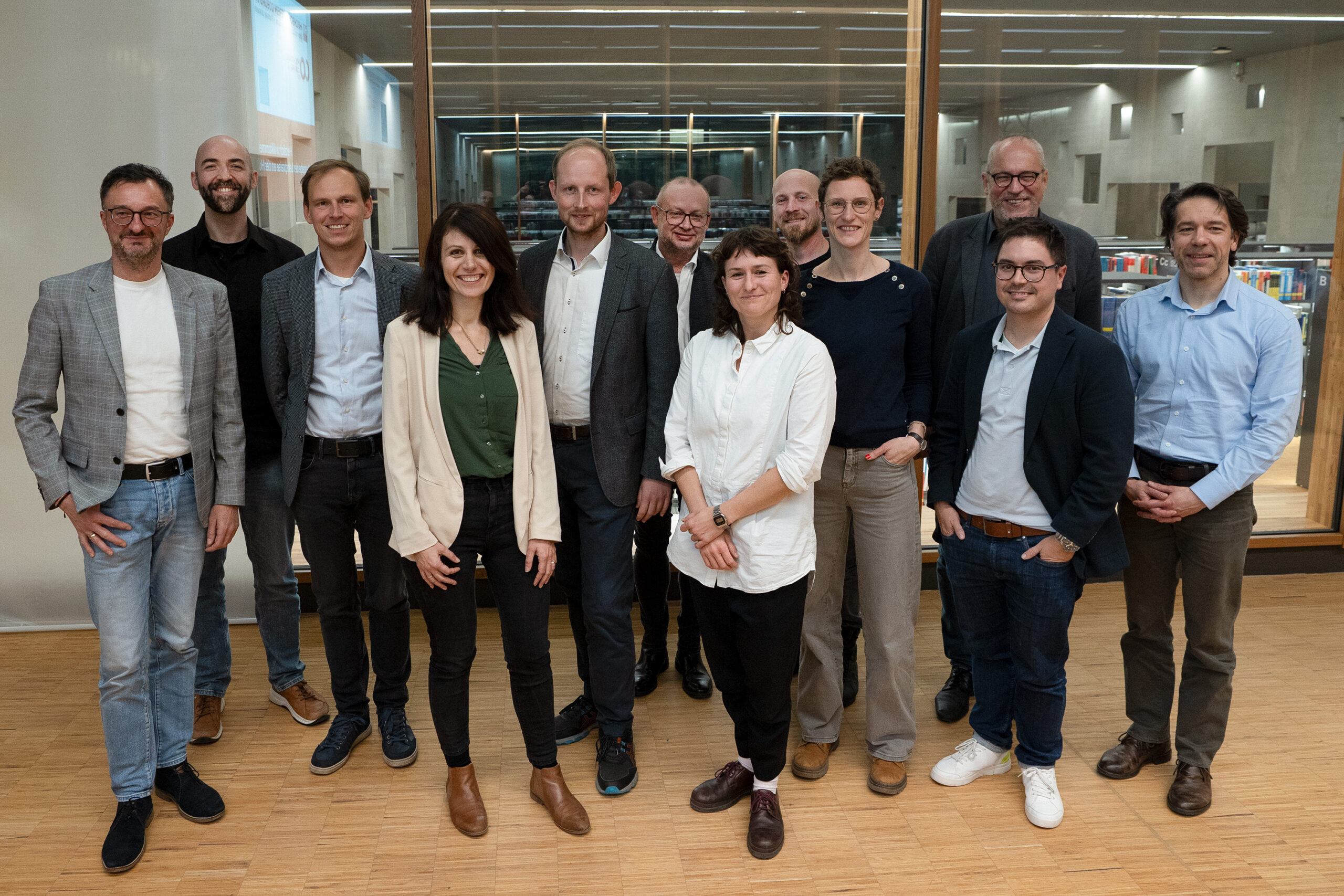 Dreizehn Personen stehen in einem Innenraum zusammen, posieren und lächeln für ein Gruppenfoto. Sie sind leger gekleidet und befinden sich in einem modernen, gut beleuchteten Raum mit Glaswänden und Holzboden. at Hochschule Coburg