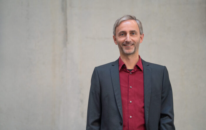 Ein Mann mit kurzen grauen Haaren und gestutztem Bart trägt eine dunkle Anzugsjacke über einem weinroten Hemd und steht lächelnd vor einem schlichten hellgrauen Hintergrund. at Hochschule Coburg