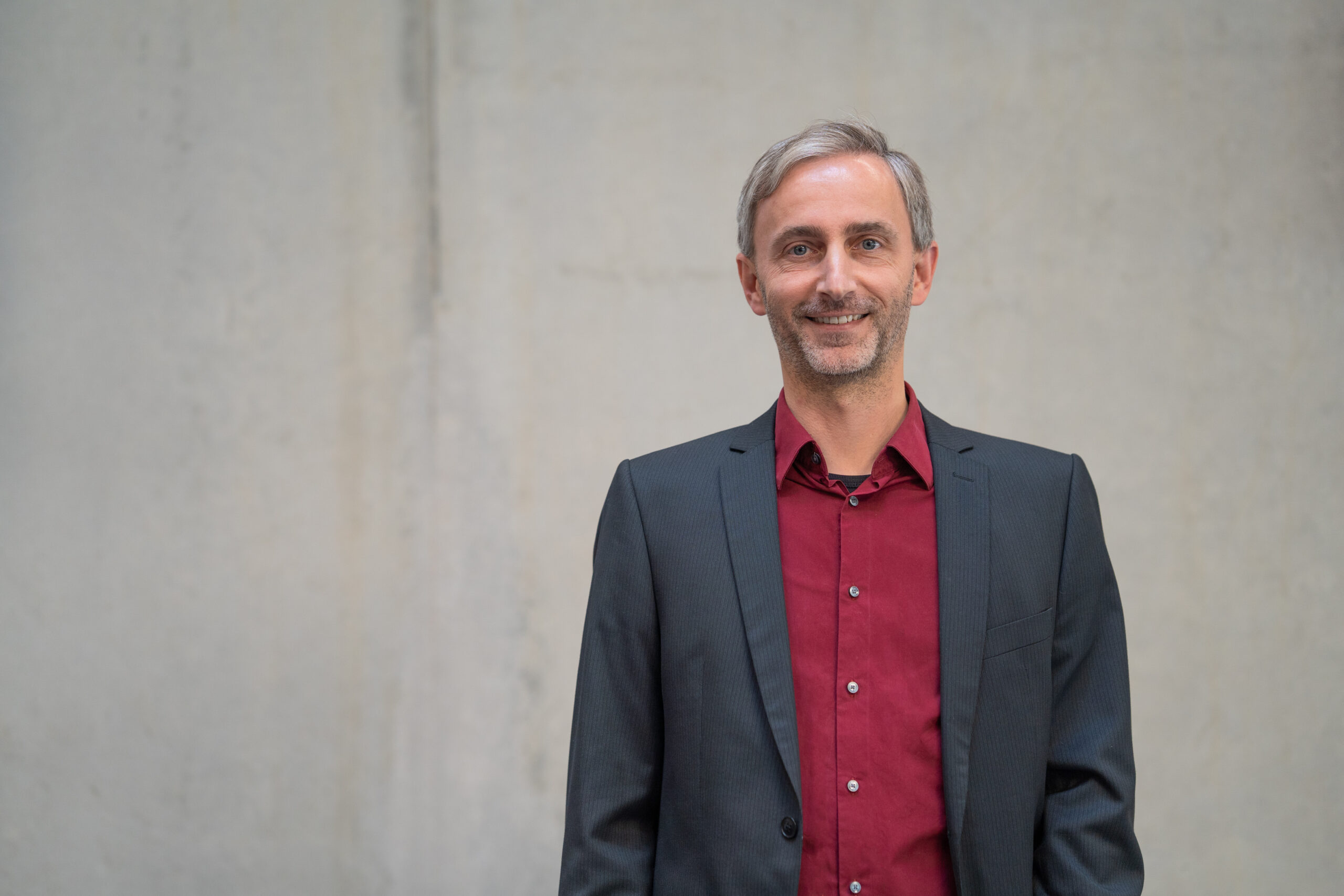 Ein Mann mit kurzen grauen Haaren und gestutztem Bart trägt eine dunkle Anzugsjacke über einem weinroten Hemd und steht lächelnd vor einem schlichten hellgrauen Hintergrund. at Hochschule Coburg