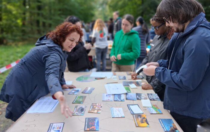 Eine Gruppe von Menschen steht um einen Tisch im Freien und beschäftigt sich mit verschiedenen Karten und Papieren zum Thema Klimabildung. Eine Frau gestikuliert, während sie spricht, und andere untersuchen Materialien in einem Waldstück in der Nähe der Hochschule. at Hochschule Coburg