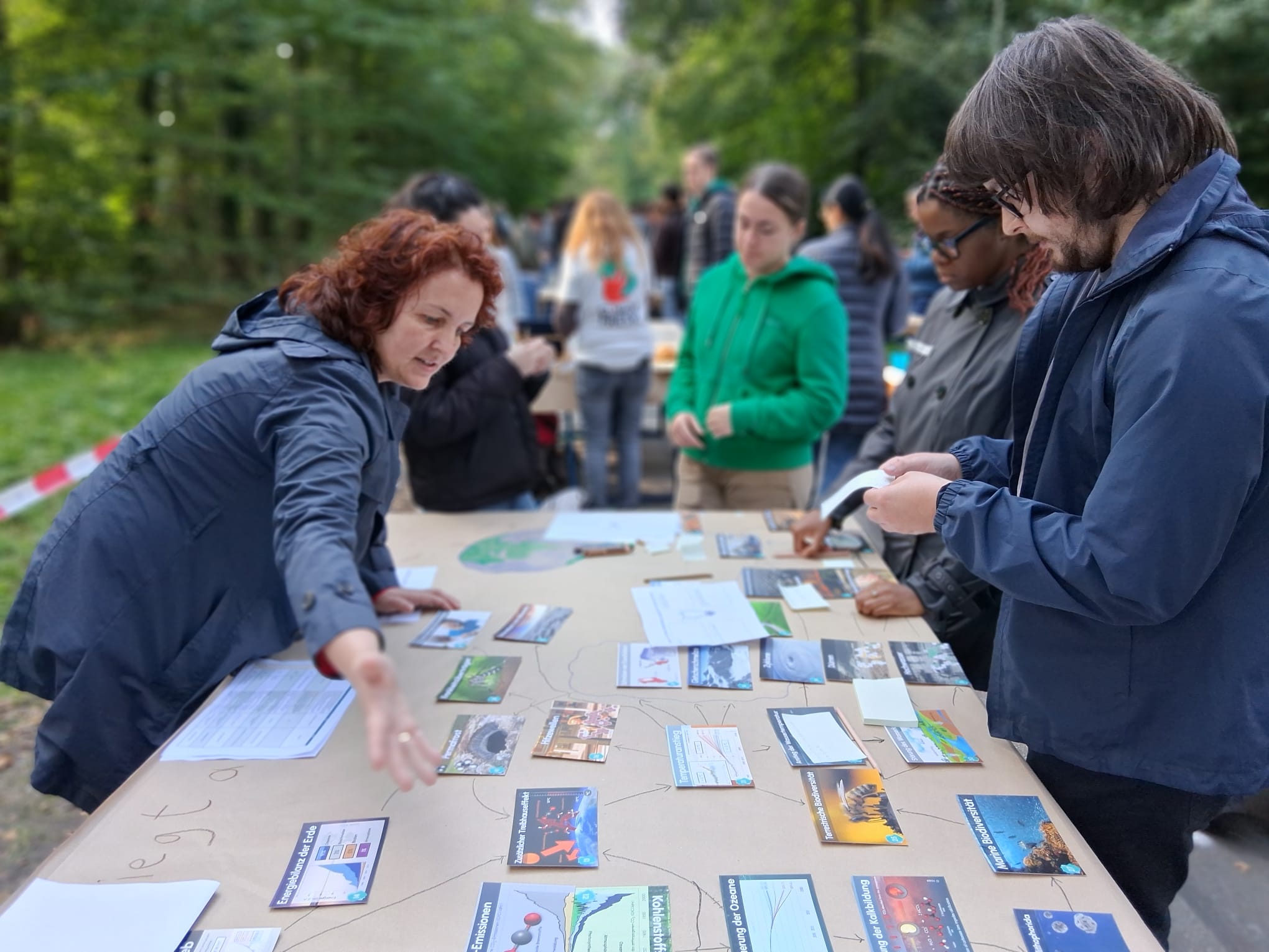 Eine Gruppe von Menschen steht um einen Tisch im Freien und beschäftigt sich mit verschiedenen Karten und Papieren zum Thema Klimabildung. Eine Frau gestikuliert, während sie spricht, und andere untersuchen Materialien in einem Waldstück in der Nähe der Hochschule. at Hochschule Coburg