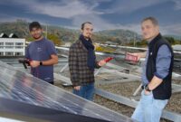 Three men stand on a rooftop next to solar panels, holding electrical testing devices. Set against city buildings and green hills, they inspect the solar installation, marking a step in their Solar Journey from Chennai to Coburg. at Hochschule Coburg