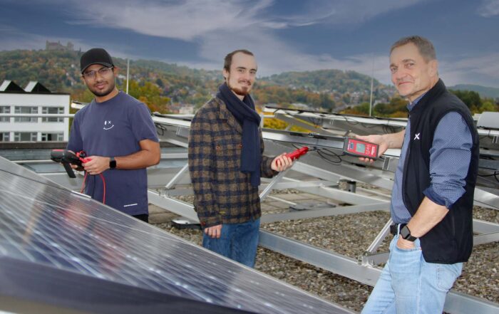 Three men stand on a rooftop next to solar panels, holding electrical testing devices. Set against city buildings and green hills, they inspect the solar installation, marking a step in their Solar Journey from Chennai to Coburg. at Hochschule Coburg