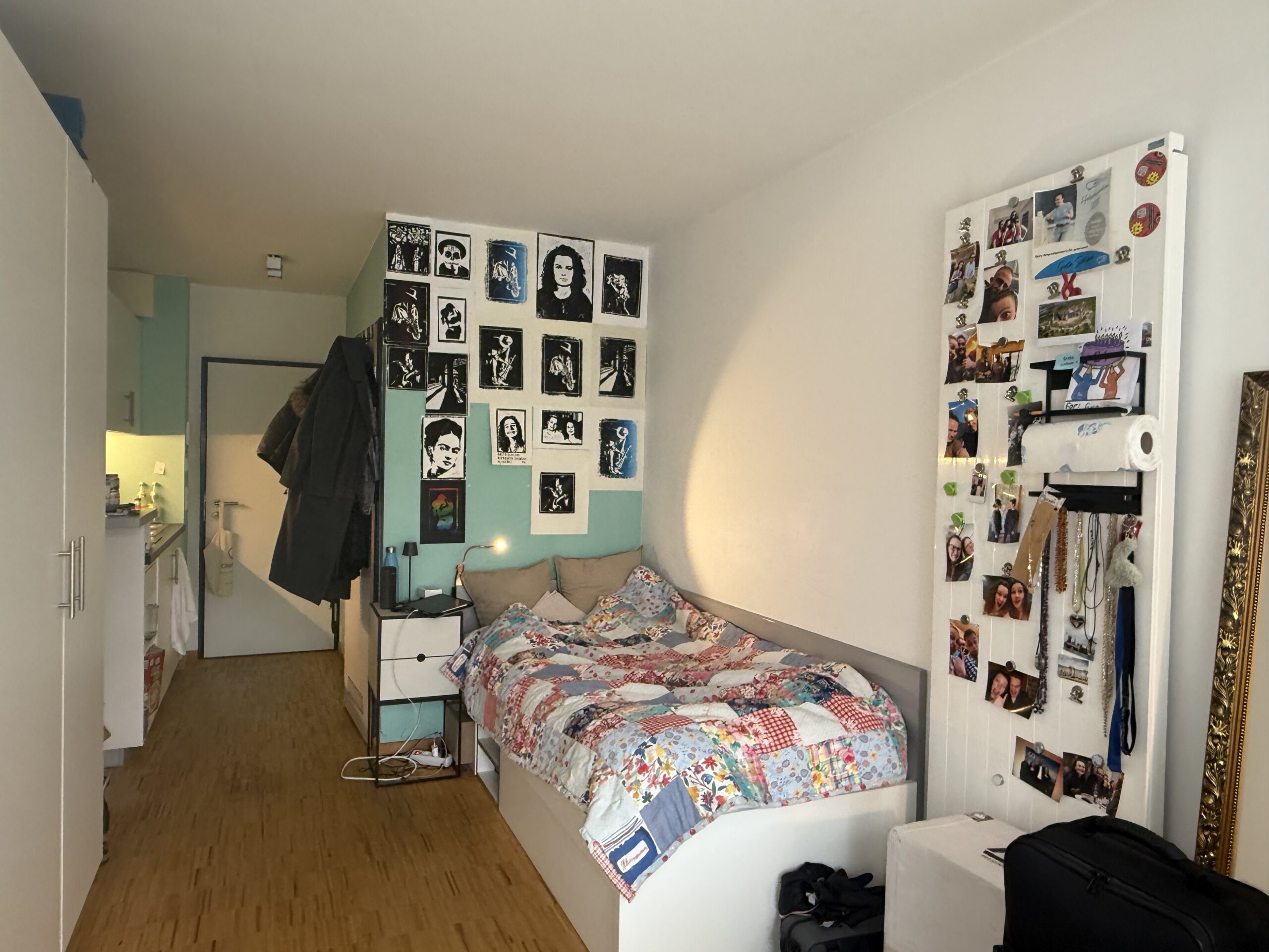 Ein gemütliches Schlafzimmer in Coburg, Bayern, mit einem Einzelbett, das mit einer Patchworkdecke bezogen ist, Schwarz-Weiß-Porträts von Prominenten über dem Bett und einer Collage aus Fotos und Souvenirs. Eine kleine Küche ist zu sehen - perfekt für das Leben in der günstigsten Hochschulstadt. at Hochschule Coburg