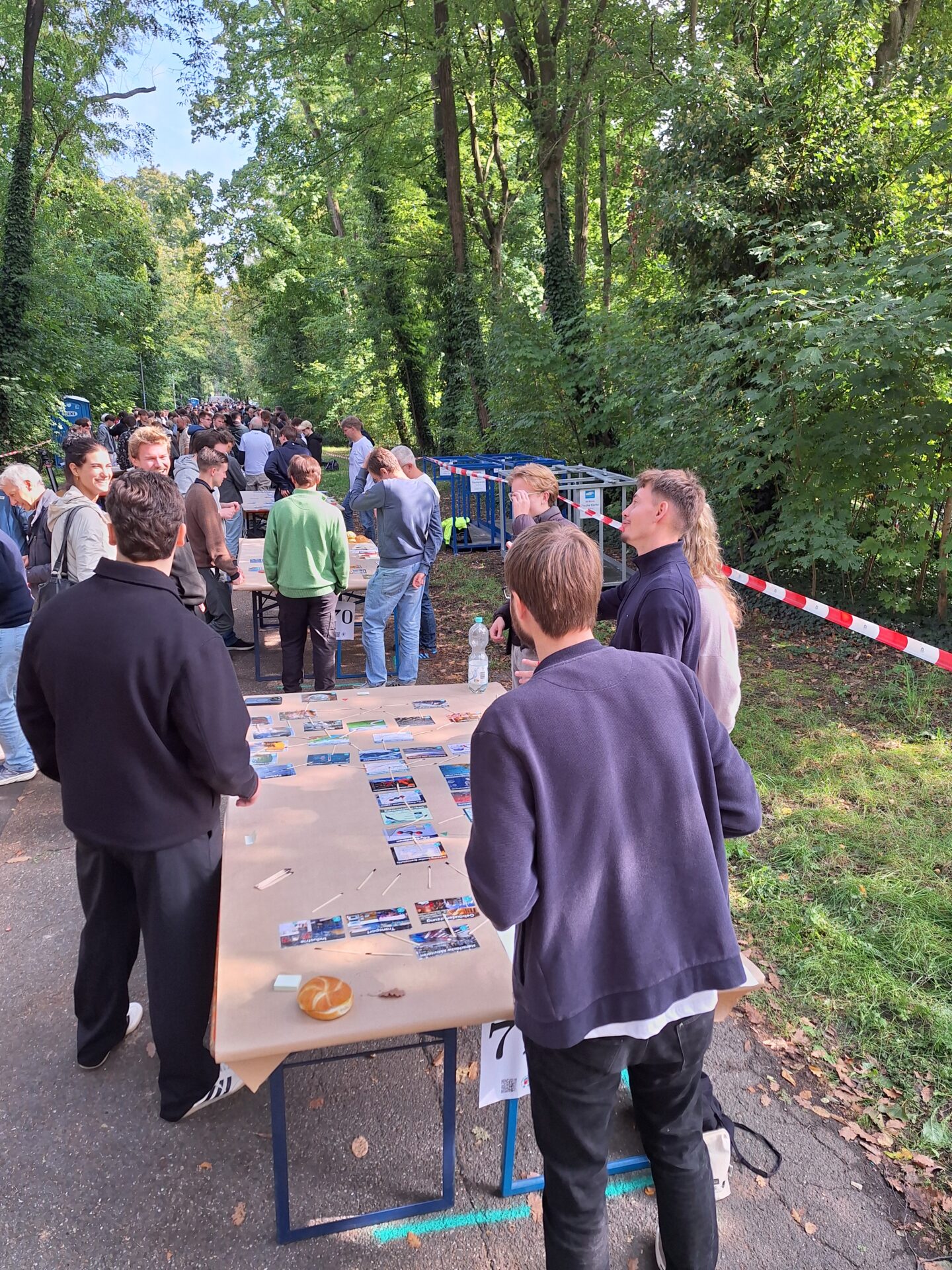Die Menschen versammeln sich im Freien an der Hochschule entlang eines Weges mit Tischen, auf denen Fotos oder Karten ausgestellt sind. Umgeben von Bäumen, Grün und rot-weißem Klebeband spiegelt die entspannte Atmosphäre ein gemeinsames Interesse an Klimabildung und der möglichen Aufstellung eines Europarekords wider. at Hochschule Coburg