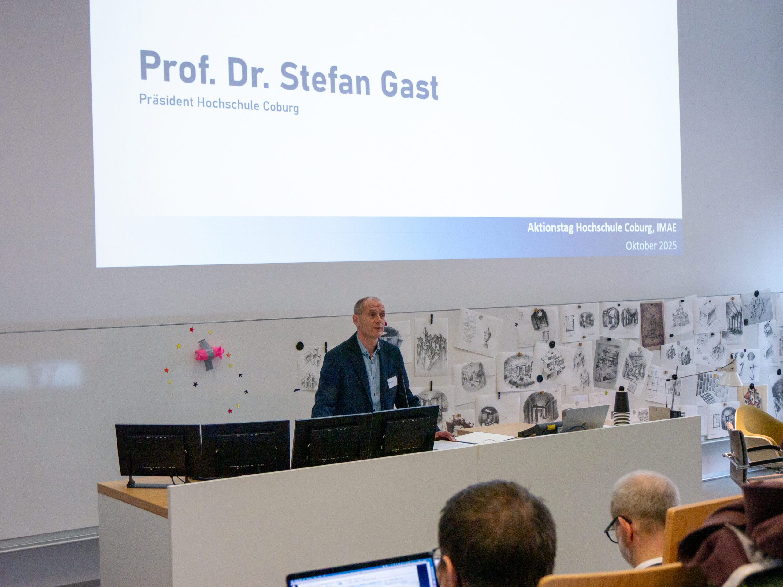 Ein Mann steht an einem Rednerpult in einem Hörsaal der Hochschule und präsentiert einen Diavortrag für den VDW-Aktionstag. Auf der Leinwand steht "Prof. Dr. Stefan Gast, Präsident Hochschule Coburg". Die Anwesenden nehmen Platz, Skizzen schmücken die Wand. at Hochschule Coburg