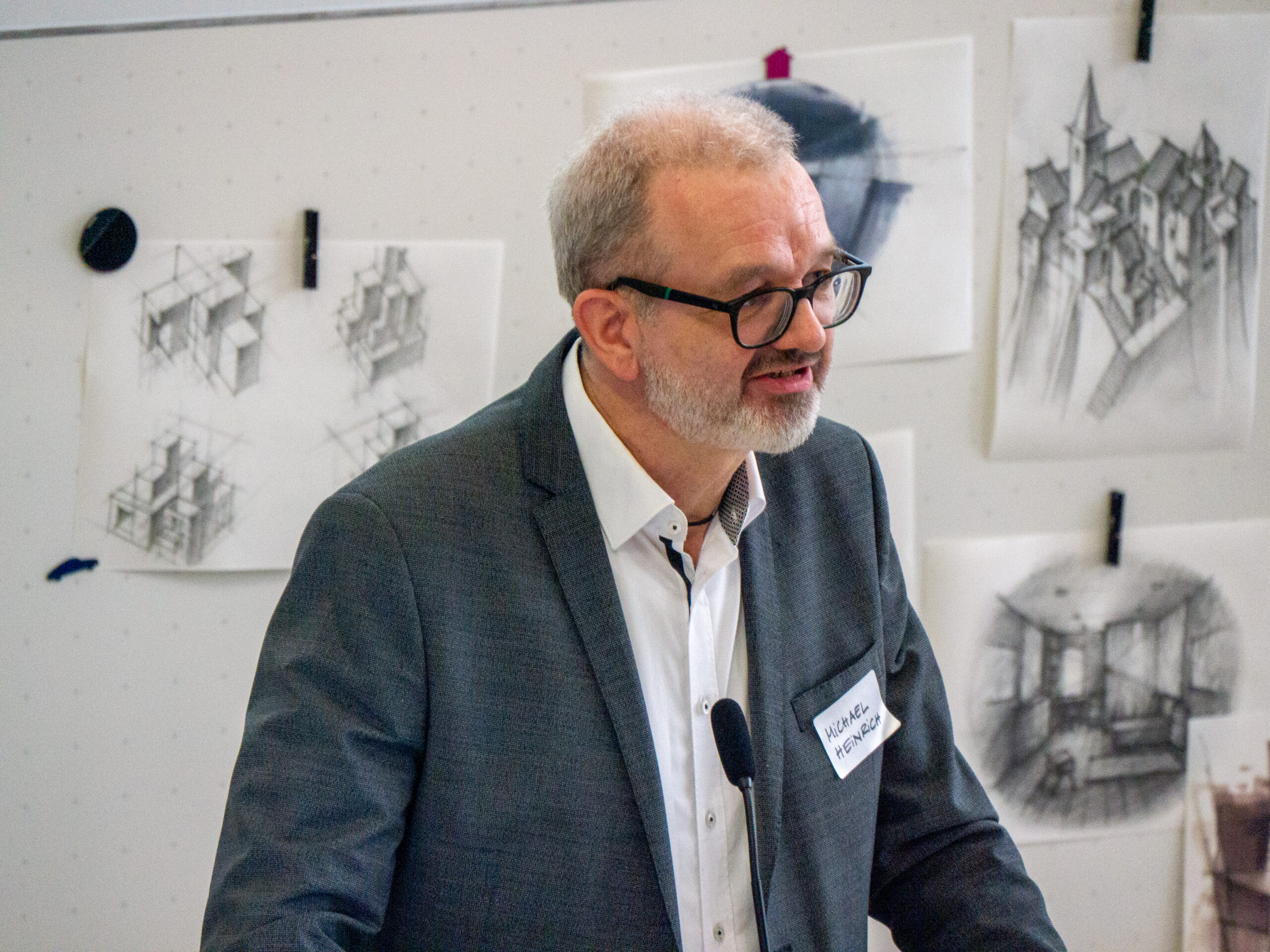 Ein Mann in einem grauen Anzug und mit Brille spricht an einem Podium in der Hochschule, während hinter ihm architektonische Skizzen von Räumen an eine Tafel geheftet sind. Er trägt ein Namensschild und scheint eine Präsentation oder eine Vorlesung zu halten. at Hochschule Coburg