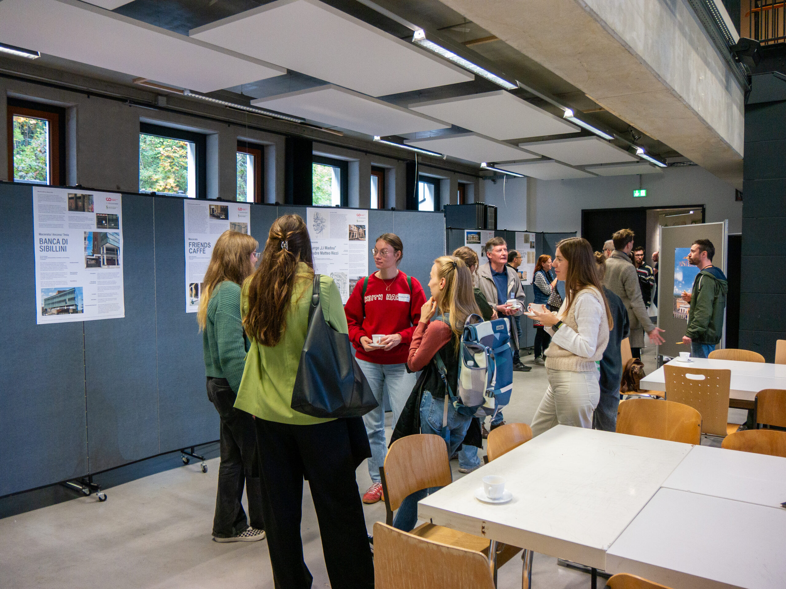 Eine Gruppe von Menschen steht während des VDW-Aktionstags in modernen Räumen der Hochschule und liest und diskutiert Plakate an Stellwänden. Einige halten Getränke in der Hand, und im Vordergrund stehen Tische mit leeren Tassen, die eine lockere, gesellige Atmosphäre schaffen. at Hochschule Coburg