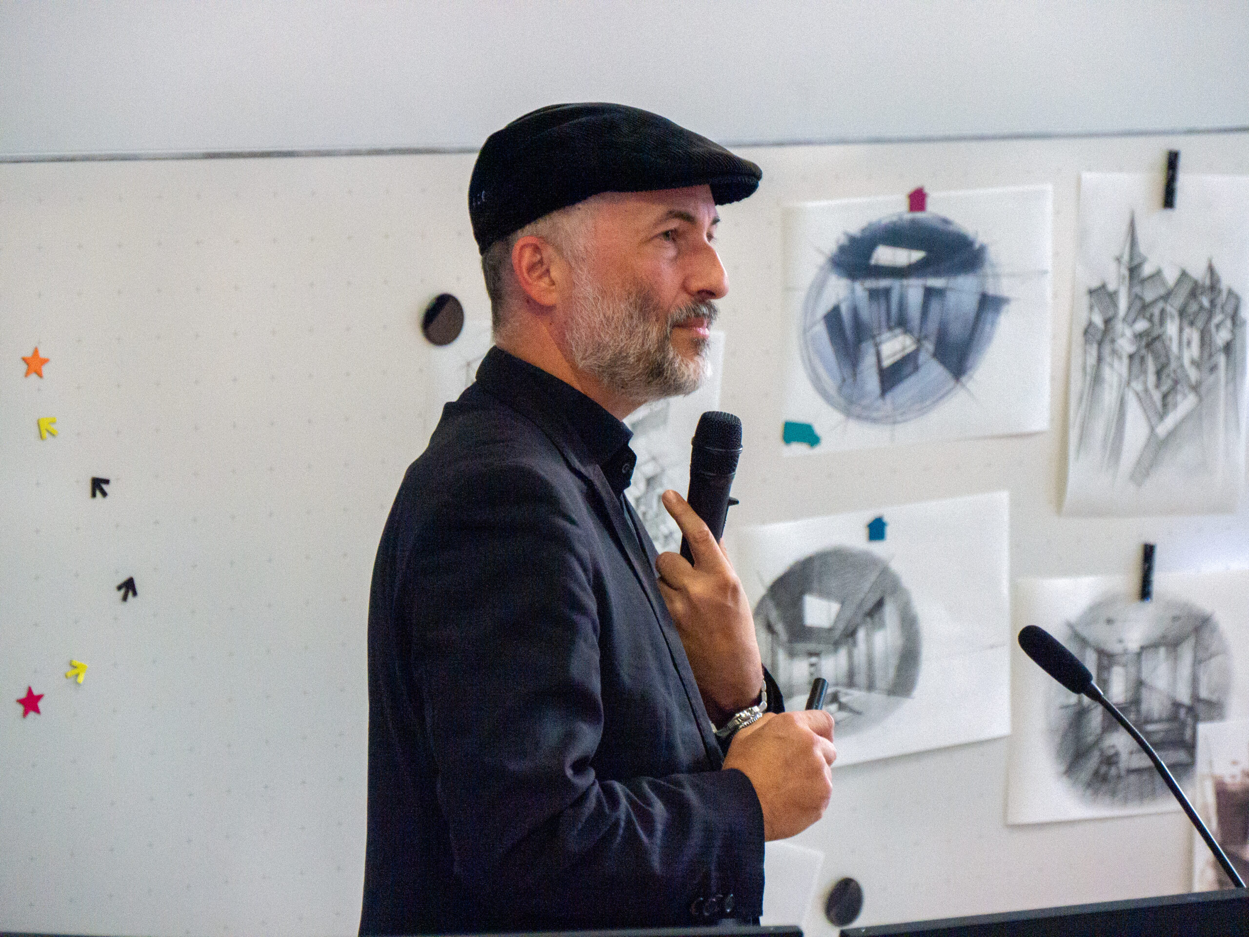 Ein Mann in dunklem Anzug und flacher Mütze hält ein Mikrofon, während er auf dem VDW-Aktionstag spricht. Hinter ihm sind architektonische Skizzen von Räumen an ein Whiteboard der Hochschule gepinnt. at Hochschule Coburg