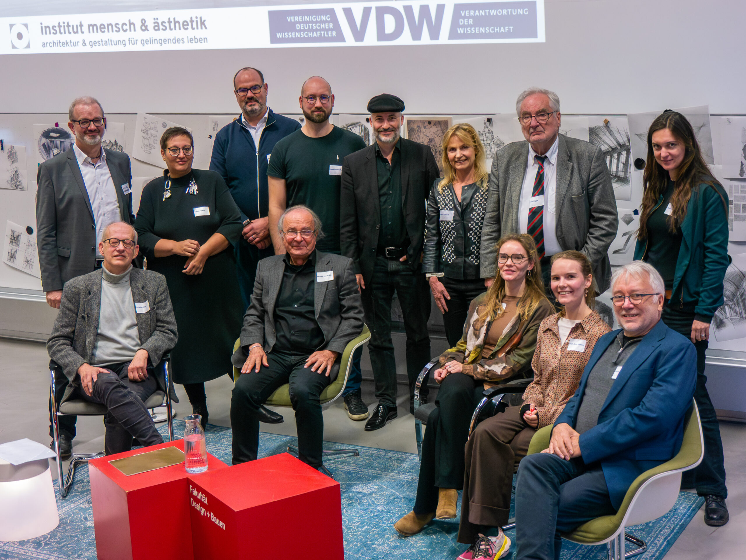 Eine Gruppe von vierzehn Erwachsenen, gekleidet in Business- und Smart-Casual-Kleidung, posiert drinnen vor einem Display mit Entwurfsskizzen und Schildern für "Institut Mensch & Ästhetik" und "VDW" während eines VDW-Aktionstages an der Hochschule Räume. Einige sitzen, andere stehen, alle lächeln. at Hochschule Coburg
