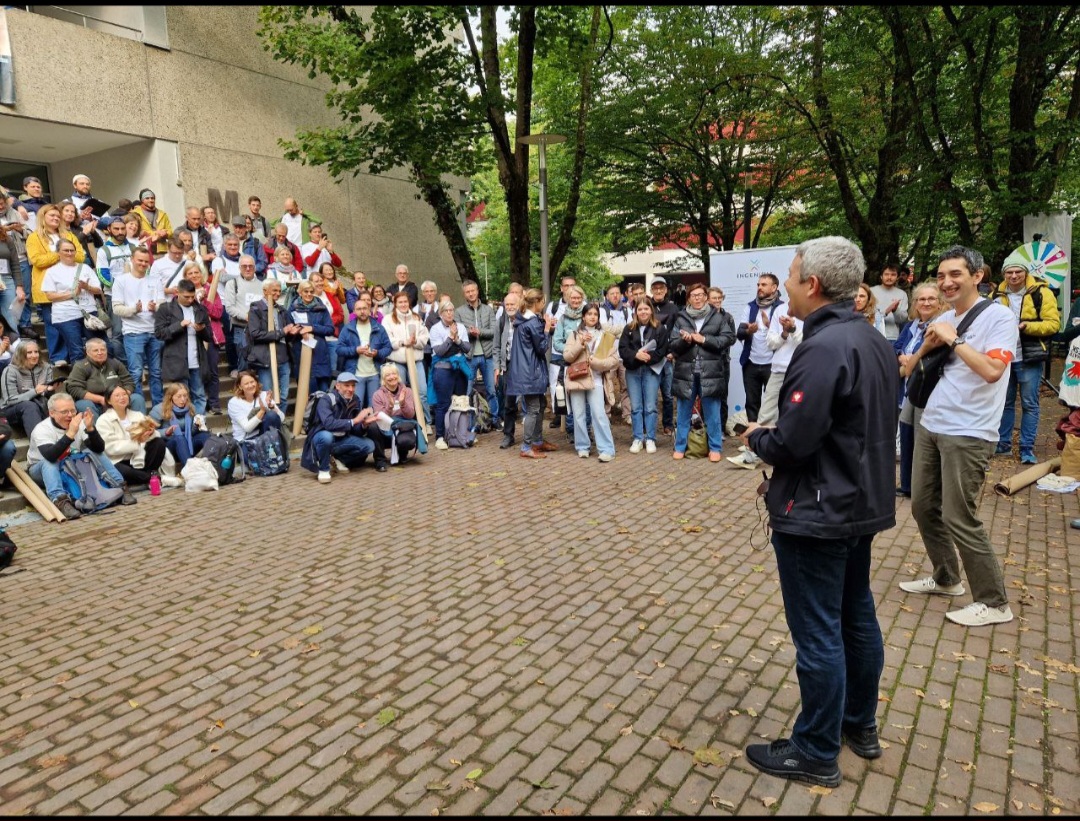 Eine große Gruppe versammelt sich im Freien auf einem Kopfsteinpflaster in der Nähe der Hochschule, einige klatschen und lächeln, während zwei Personen im Vordergrund stehen und in die Menge schauen. Im Hintergrund sind Bäume und Gebäude bei dieser Klimabildung Europarekord zu sehen. at Hochschule Coburg