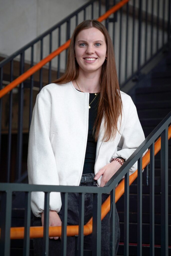 A young woman with long brown hair, wearing a white jacket over a black top, stands smiling next to a staircase with black railings and orange handrails. at Hochschule Coburg
