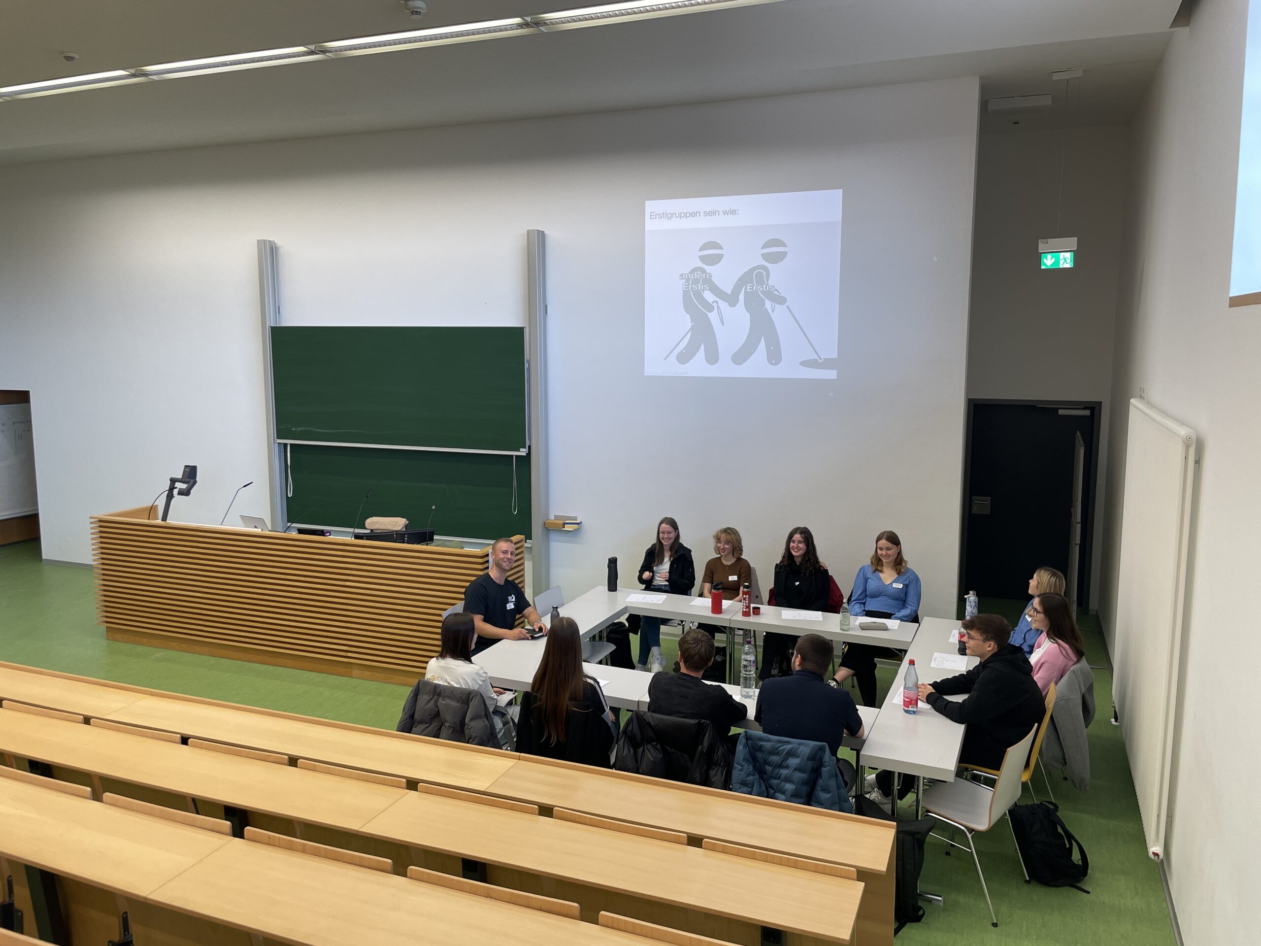 Eine Gruppe von Studenten, wahrscheinlich Erstsemester bei ihrem Studienstart, sitzt an Tischen in einem Universitätshörsaal und blickt auf einen Vortragenden. Hinter ihnen befindet sich eine Kreidetafel und eine projizierte Folie mit Cartoon-Figuren, mit leeren Sitzreihen im Vordergrund. at Hochschule Coburg