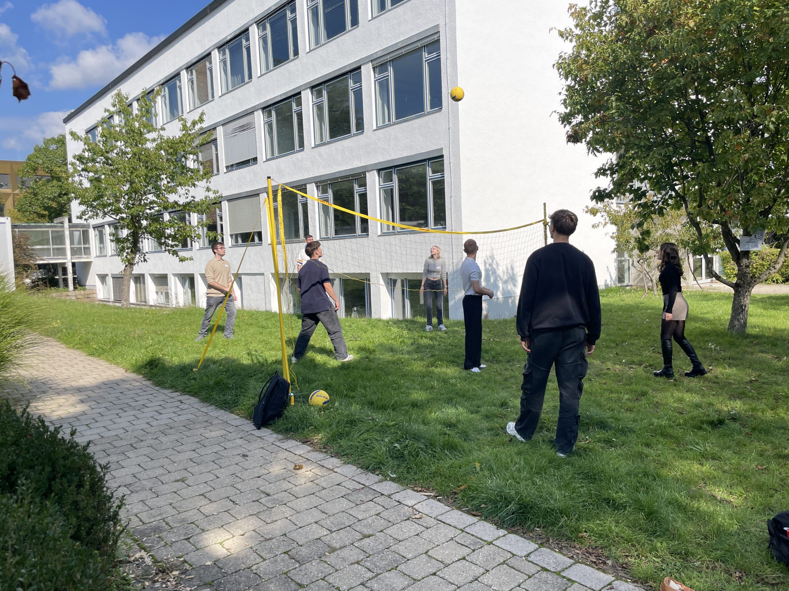 Sechs Personen spielen an den Erstsemestertagen auf einer Wiese vor einem modernen, weißen Gebäude Volleyball - eine perfekte Gelegenheit, gemeinsam zu starten und gut anzukommen. Das Wetter ist sonnig mit ein paar Wolken und Bäumen in der Nähe. at Hochschule Coburg