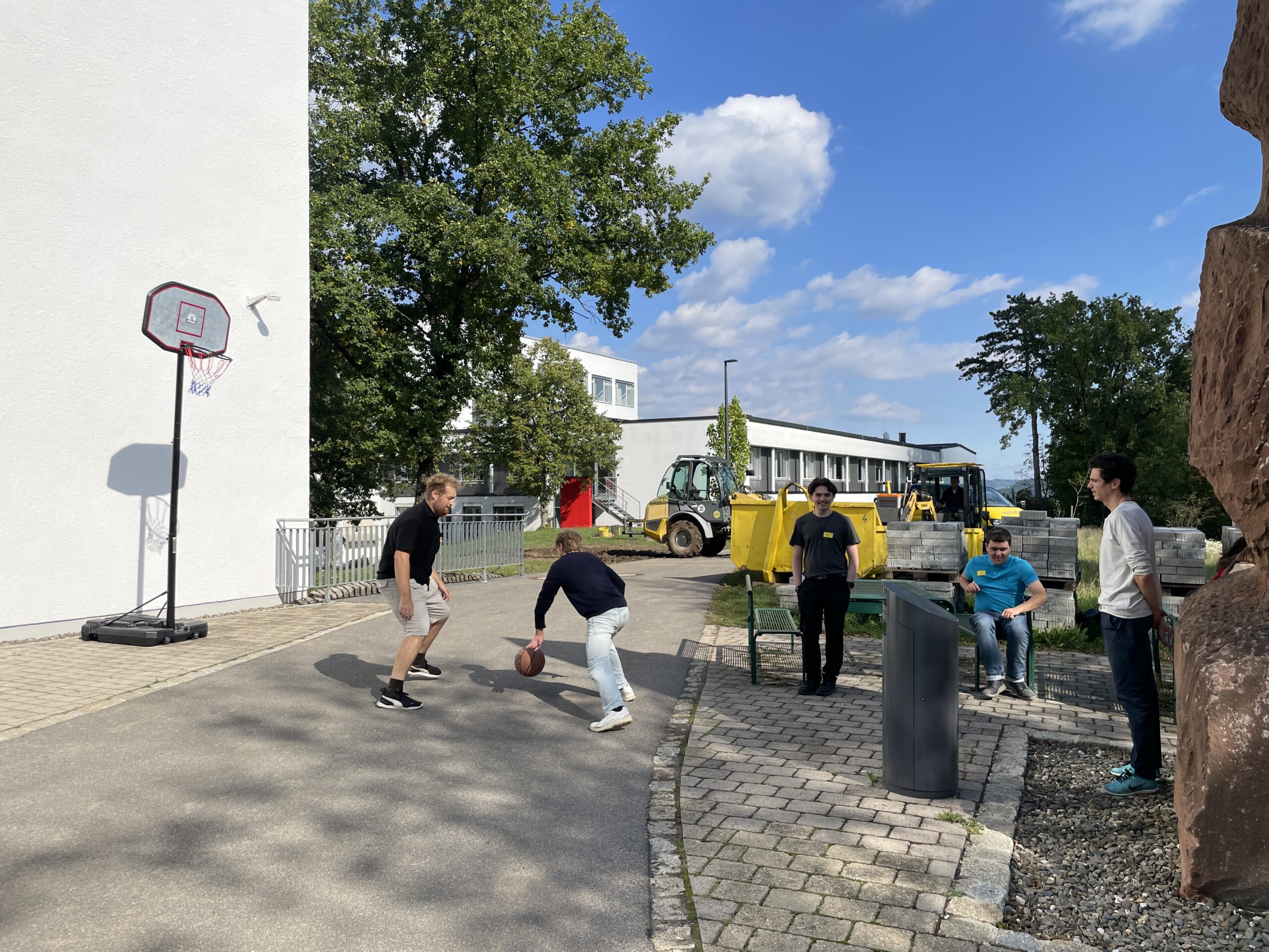 Zwei Personen spielen Basketball auf einem Platz im Freien in der Nähe eines weißen Gebäudes, während drei andere zusehen - perfekt für Erstsemestertage zum gemeinsamen Start. Im Hintergrund sind bei teilweise bewölktem Himmel Baufahrzeuge und -geräte zu sehen. at Hochschule Coburg