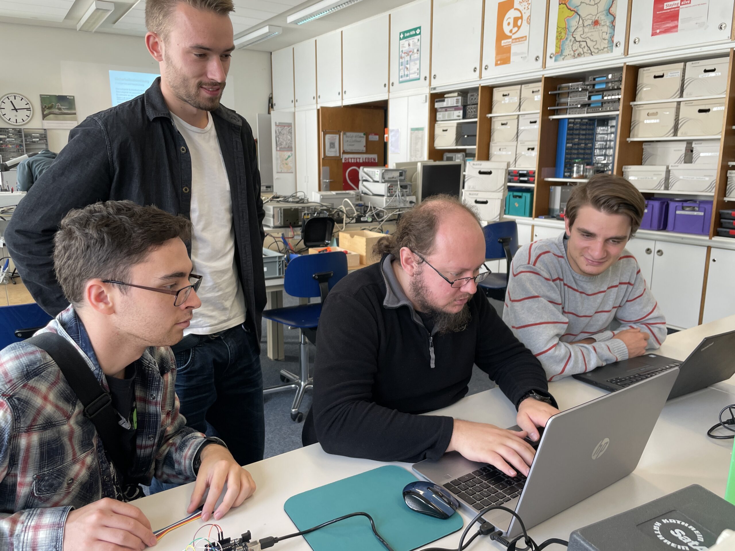 Vier Männer arbeiten zusammen in einem Klassenzimmer oder Labor, diskutieren und tippen auf Laptops. Im Hintergrund sind elektronische Geräte und Lagerregale zu sehen, während sie gemeinsam starten und sicherstellen, dass alle gut ankommen. at Hochschule Coburg