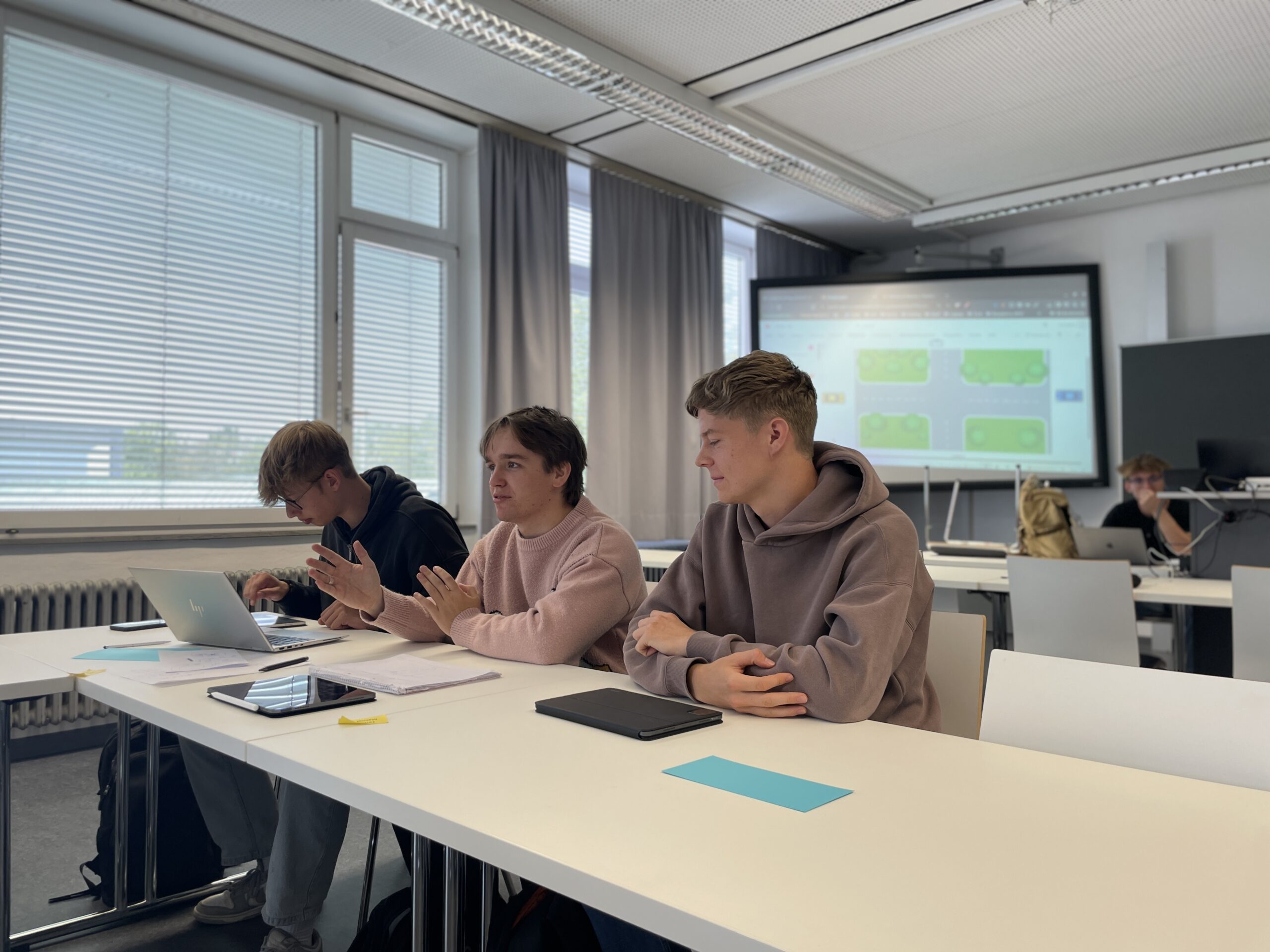 Drei junge Männer sitzen mit Laptops und Papieren an einem Tisch in einem Klassenzimmer und diskutieren während ihres Studienstarts. Auf einem großen Bildschirm wird eine Präsentation gezeigt, und weiter hinten sind andere Studenten zu sehen. at Hochschule Coburg