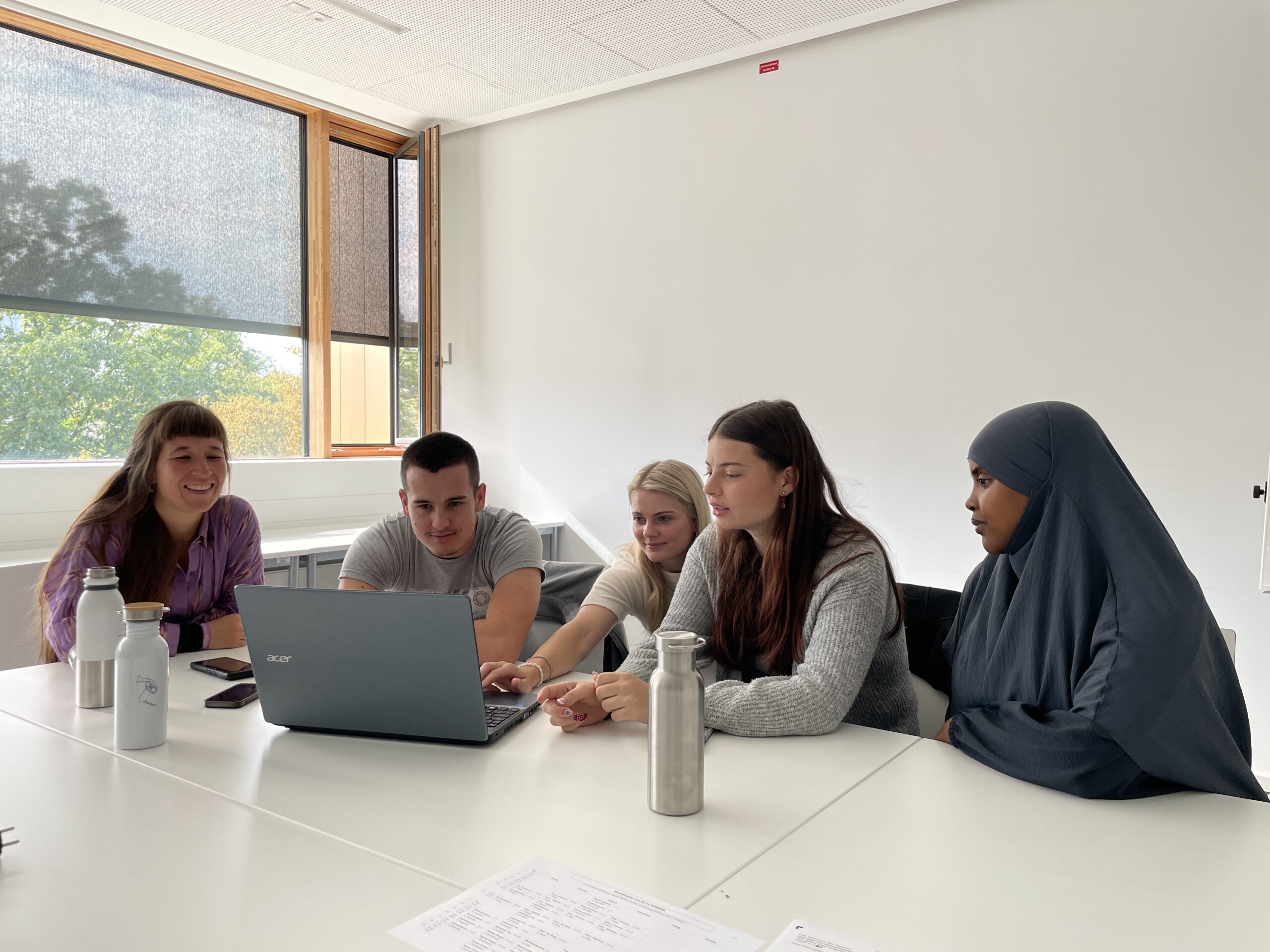 Fünf Personen sitzen in einem hellen Raum um einen Tisch und schauen auf einen Laptop. Drei Frauen und ein Mann sind beschäftigt, während eine weitere Frau im Hidschab beobachtet - ein idealer Moment, um gemeinsam zu starten und bei den Erstsemestertagen gut anzukommen. at Hochschule Coburg