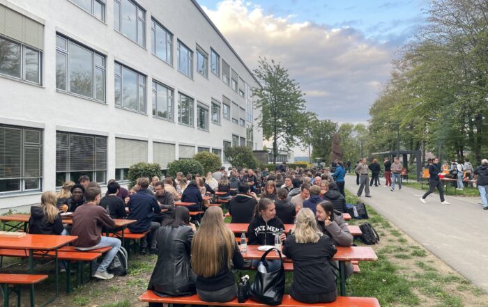 Während der Erstsemestertage sitzt eine große Gruppe an langen orangefarbenen Picknicktischen vor einem weißen Gebäude, um Kontakte zu knüpfen und zu essen. Umgeben von Bäumen und Wegen, unter einem teilweise bewölkten Himmel, können alle gemeinsam starten und gut ankommen. at Hochschule Coburg