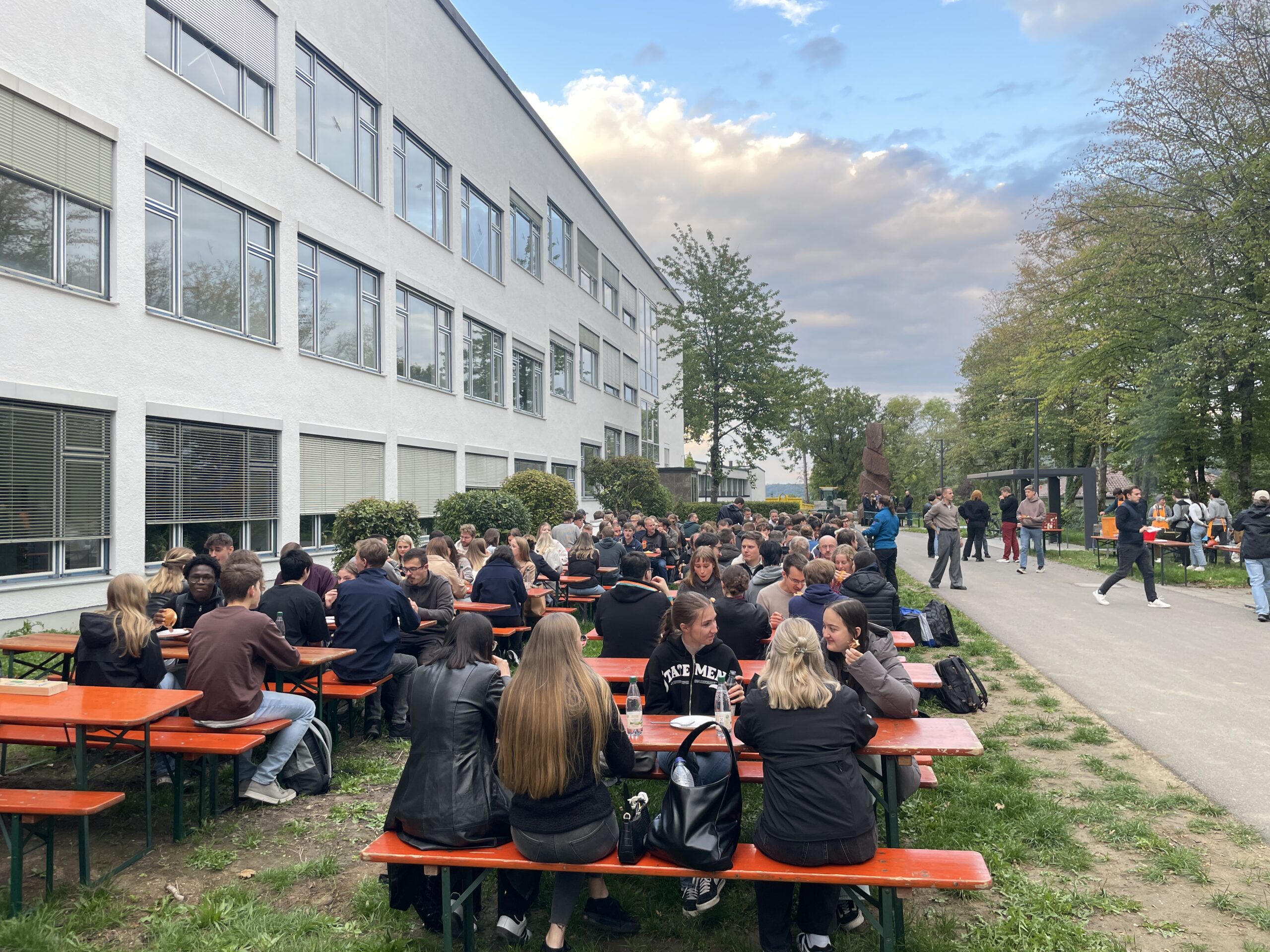 Während der Erstsemestertage sitzt eine große Gruppe an langen orangefarbenen Picknicktischen vor einem weißen Gebäude, um Kontakte zu knüpfen und zu essen. Umgeben von Bäumen und Wegen, unter einem teilweise bewölkten Himmel, können alle gemeinsam starten und gut ankommen. at Hochschule Coburg