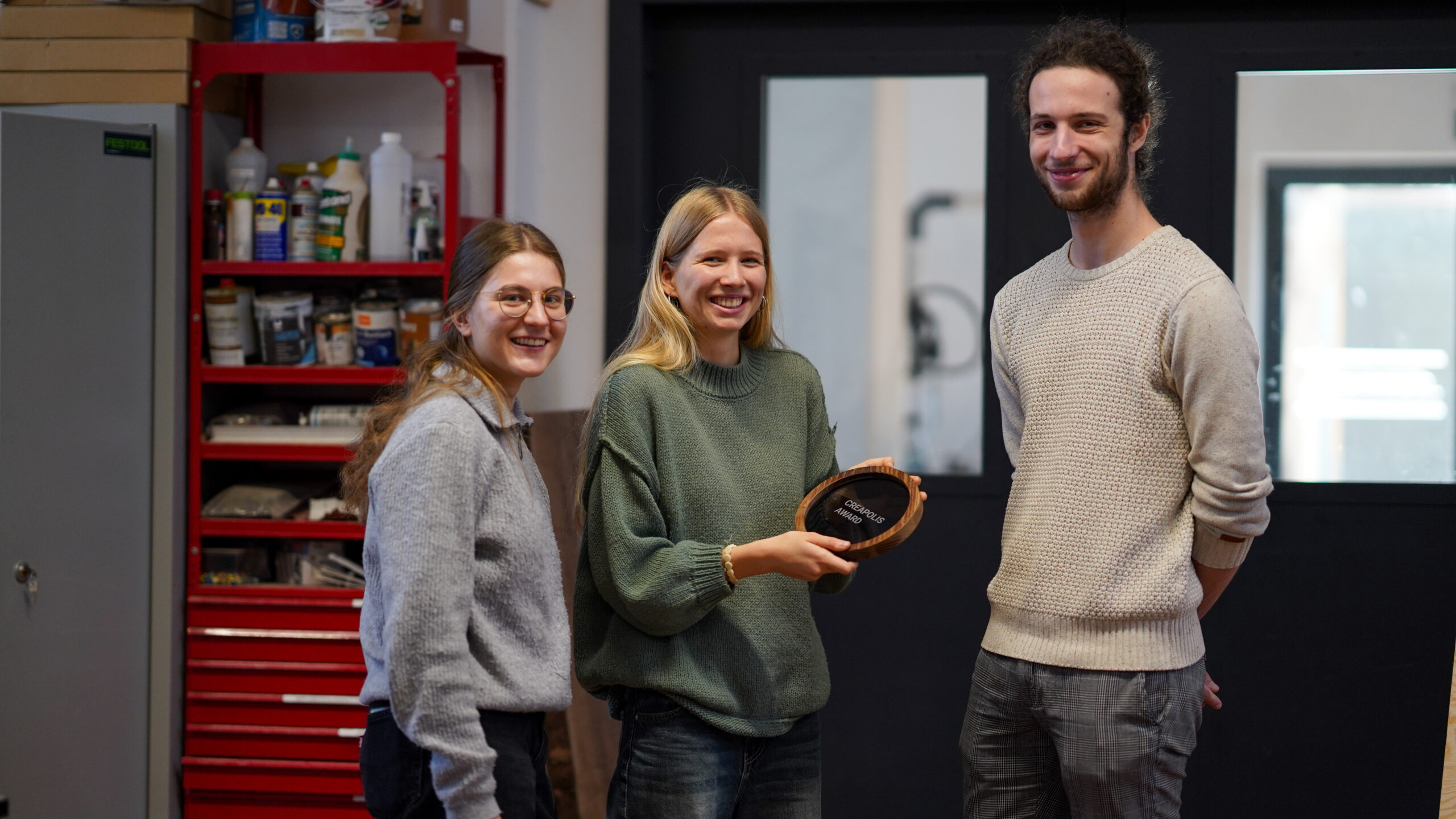 Drei Personen stehen in einem Haus und lächeln in die Kamera. Die Person in der Mitte hält einen holzgerahmten CREAPOLIS-Preis, mit dem ihre Innovation gewürdigt wird. Im Hintergrund sind Regale mit Werkzeugen und Zubehör zu sehen. at Hochschule Coburg