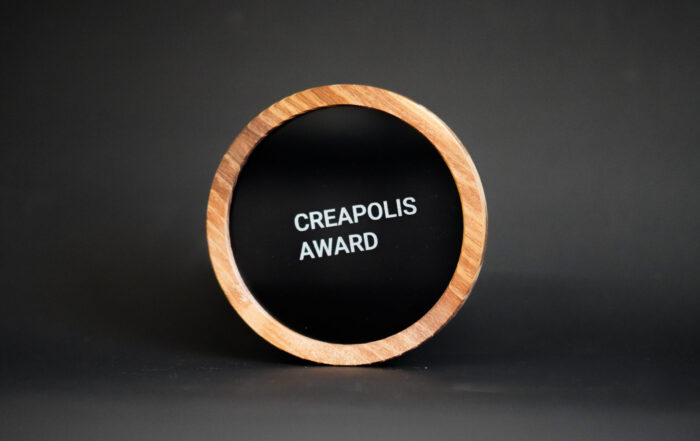 Eine runde, holzgerahmte Plakette mit schwarzer Mitte, auf der in weißer Schrift der Text "CREAPOLIS AWARD" steht, der Innovation und Design auszeichnet, vor einem schlichten dunklen Hintergrund. at Hochschule Coburg