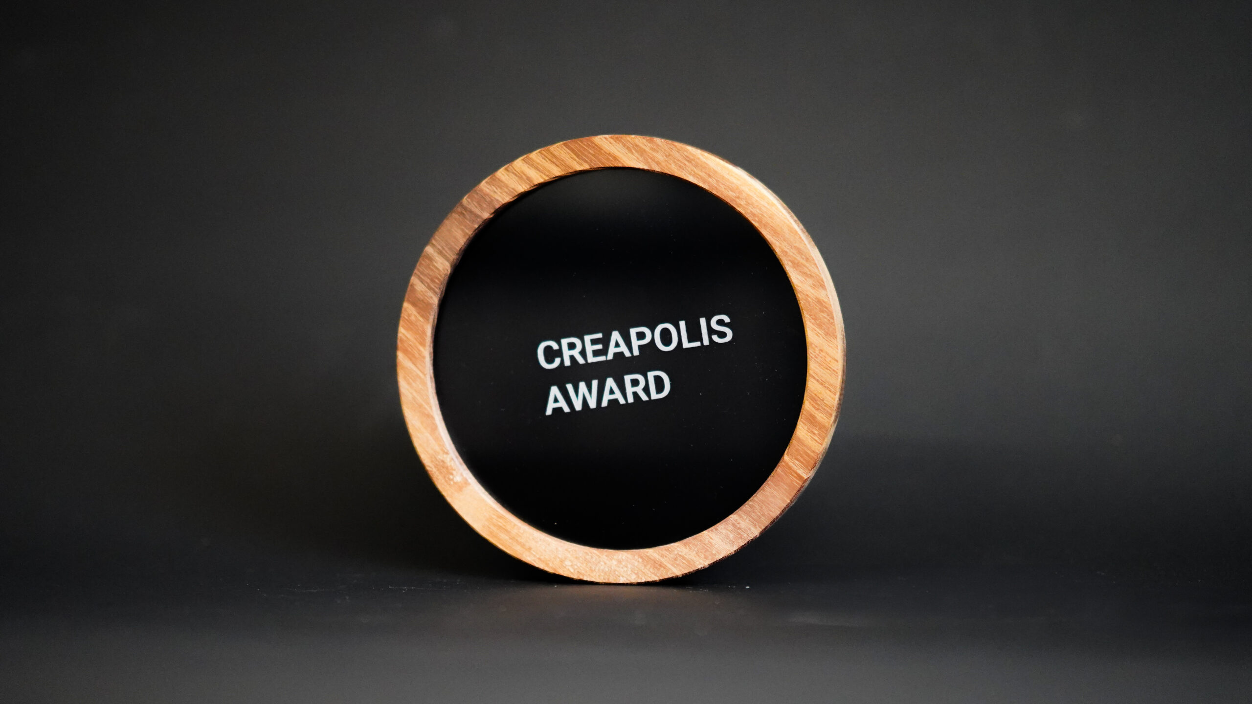 Eine runde, holzgerahmte Plakette mit schwarzer Mitte, auf der in weißer Schrift der Text "CREAPOLIS AWARD" steht, der Innovation und Design auszeichnet, vor einem schlichten dunklen Hintergrund. at Hochschule Coburg
