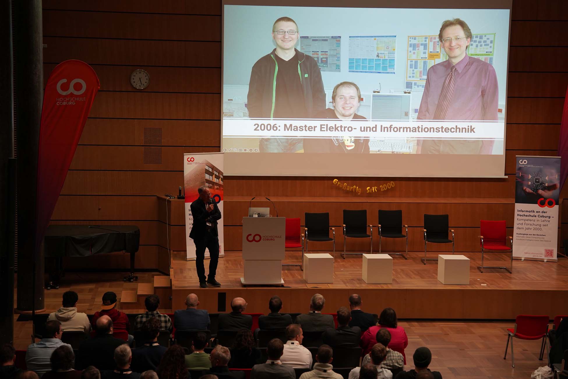 Ein Referent hält in einem Hörsaal einen Vortrag vor Publikum, dahinter zeigt eine große Leinwand ein Foto von drei lächelnden Menschen mit der Aufschrift "2006: Master Elektro- und Informationstechnik" bei der Veranstaltung Informatik und KI der Hochschule Coburg. at Hochschule Coburg