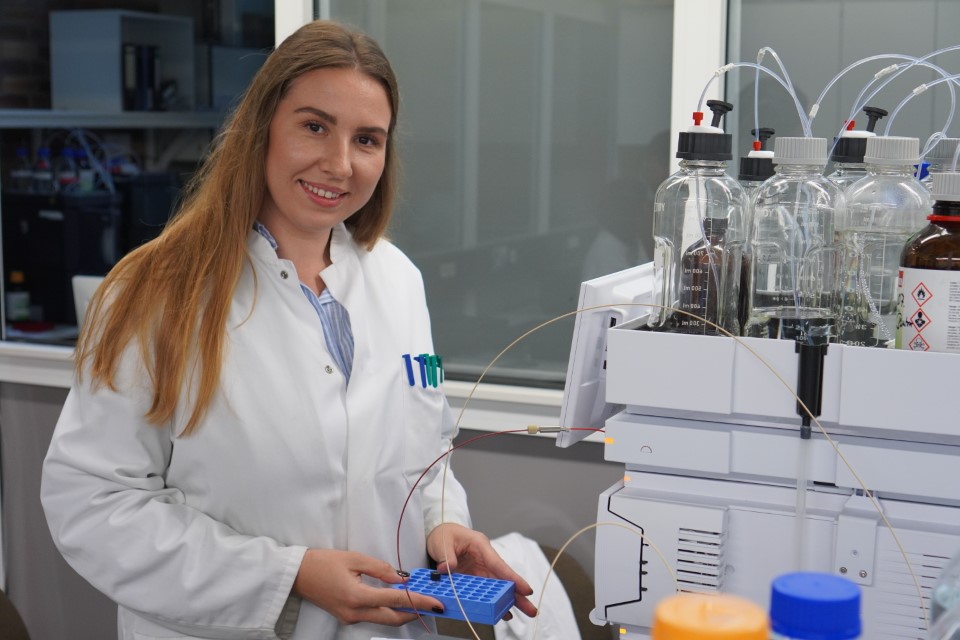 Eine Frau im Laborkittel lächelt, während sie ein blaues Reagenzglasgestell in einem Labor der Hochschule hält, umgeben von wissenschaftlichen Geräten und Flaschen - sie forscht an einer neuen Therapie gegen Krebs. at Hochschule Coburg