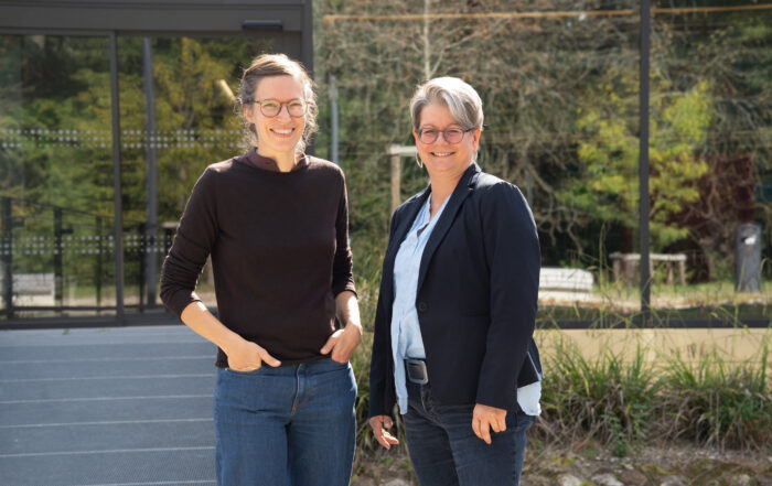 Zwei Frauen stehen im Freien vor einem Glasgebäude und lächeln in die Kamera. Die eine trägt einen schwarzen Pullover und Jeans, die andere einen Blazer über einer hellblauen Bluse und Jeans, um Teamgeist zu zeigen. Im Hintergrund sind Bäume und Grünflächen zu sehen. at Hochschule Coburg