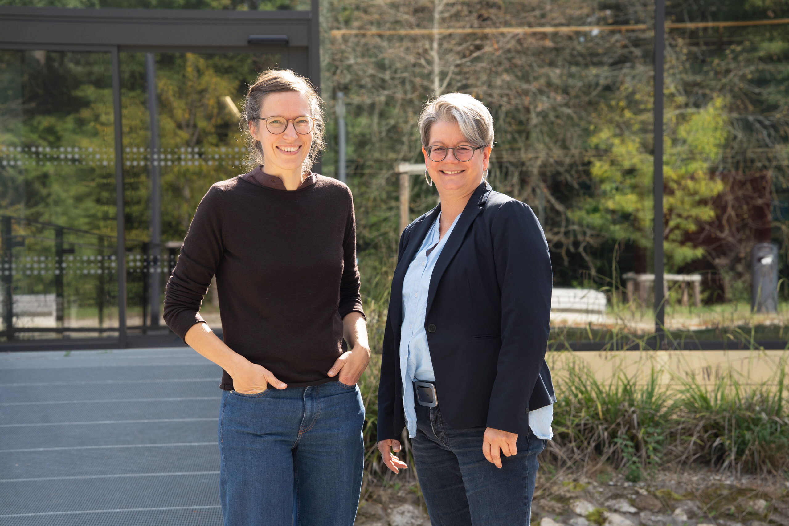 Zwei Frauen stehen im Freien vor einem Glasgebäude und lächeln in die Kamera. Die eine trägt einen schwarzen Pullover und Jeans, die andere einen Blazer über einer hellblauen Bluse und Jeans, um Teamgeist zu zeigen. Im Hintergrund sind Bäume und Grünflächen zu sehen. at Hochschule Coburg