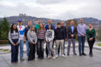 A group of thirteen people, both men and women, stand outdoors on a paved surface with a cityscape and hills in the background. The sky is cloudy, and everyone is casually dressed, smiling at the camera. at Hochschule Coburg