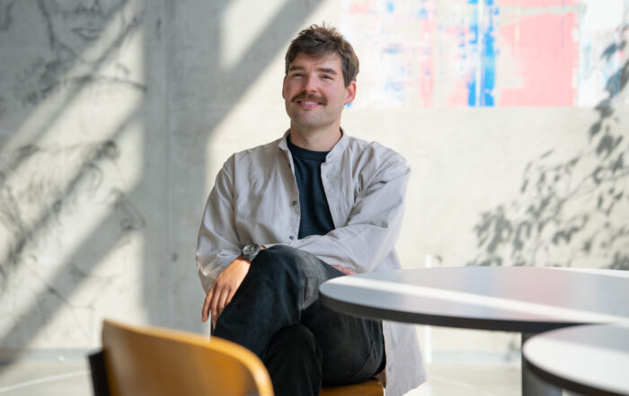 Prof. Buchmüller, mit Schnurrbart, hellem Hemd und dunkler Hose, sitzt im Schneidersitz auf einem Stuhl an einem runden Tisch in einem hellen, modernen Raum mit abstrakter Wandkunst und natürlichem Licht. at Hochschule Coburg