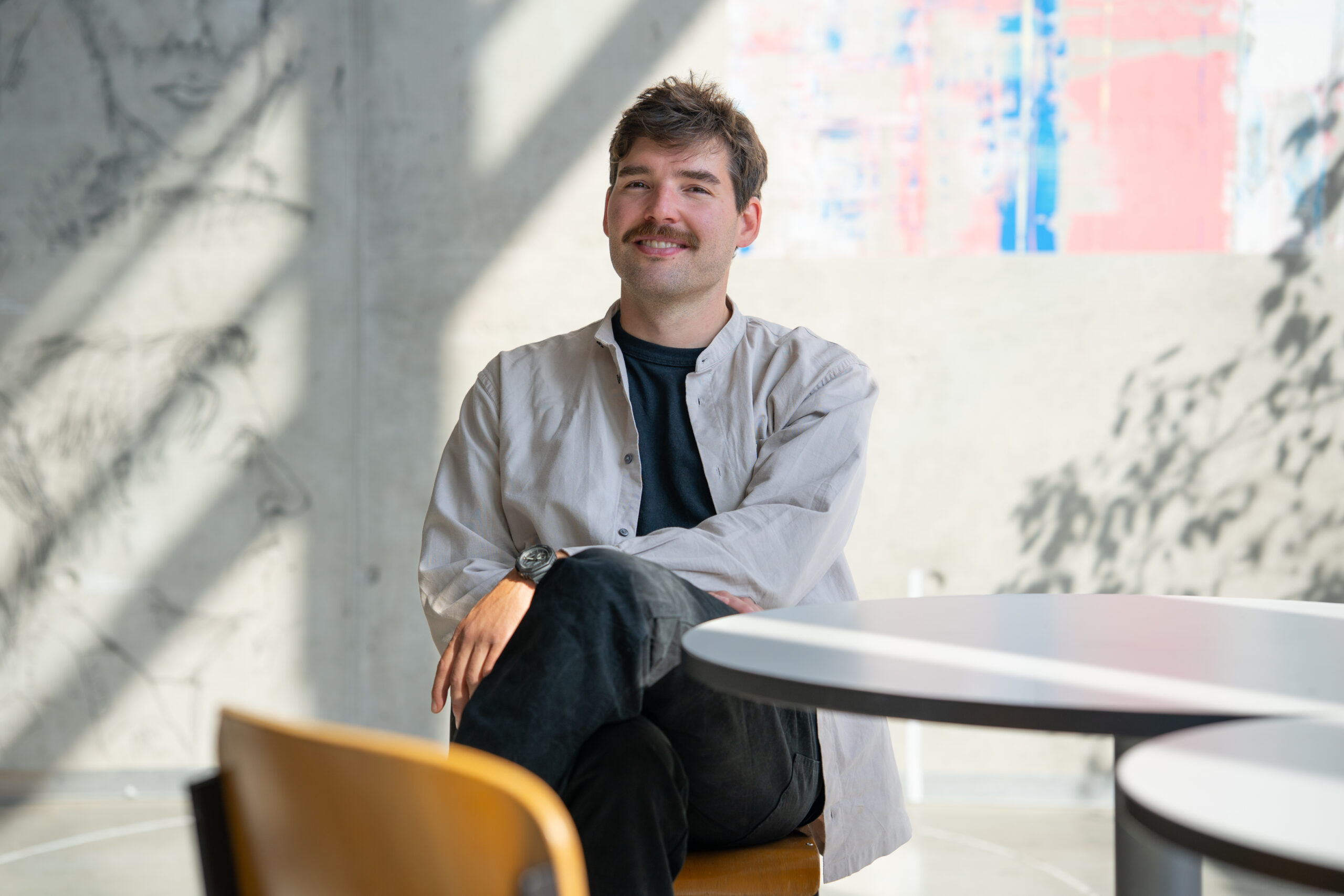 Prof. Buchmüller, mit Schnurrbart, hellem Hemd und dunkler Hose, sitzt im Schneidersitz auf einem Stuhl an einem runden Tisch in einem hellen, modernen Raum mit abstrakter Wandkunst und natürlichem Licht. at Hochschule Coburg