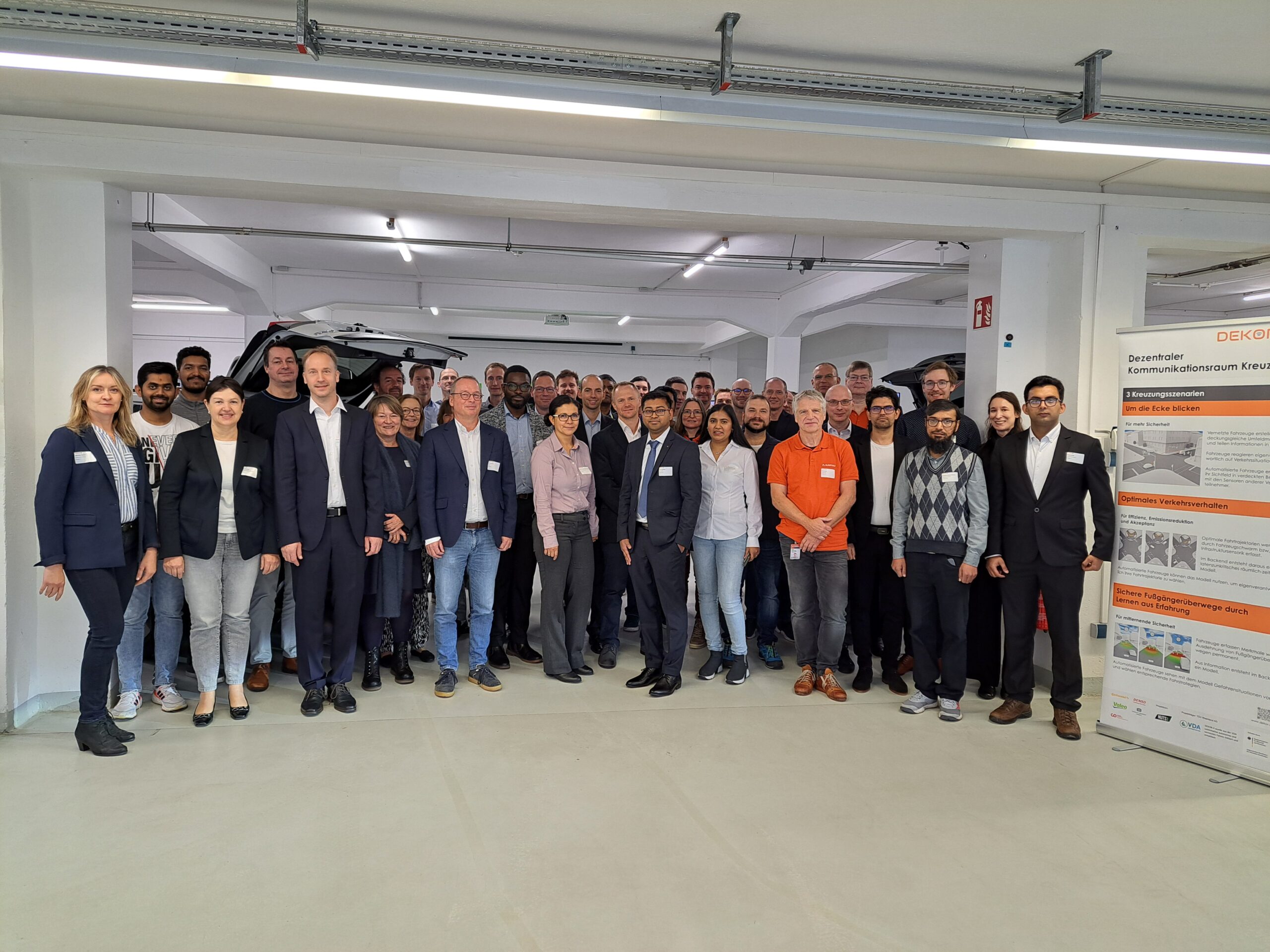 Eine große Gruppe von professionell gekleideten Menschen steht in einem Innenraum und lächelt in die Kamera. Vor einer weißen Säule versammelt, mit DEKOR-X-Informationsplakaten auf der rechten Seite, werben sie für smarte Kommunikation für sichere Straßen. at Hochschule Coburg