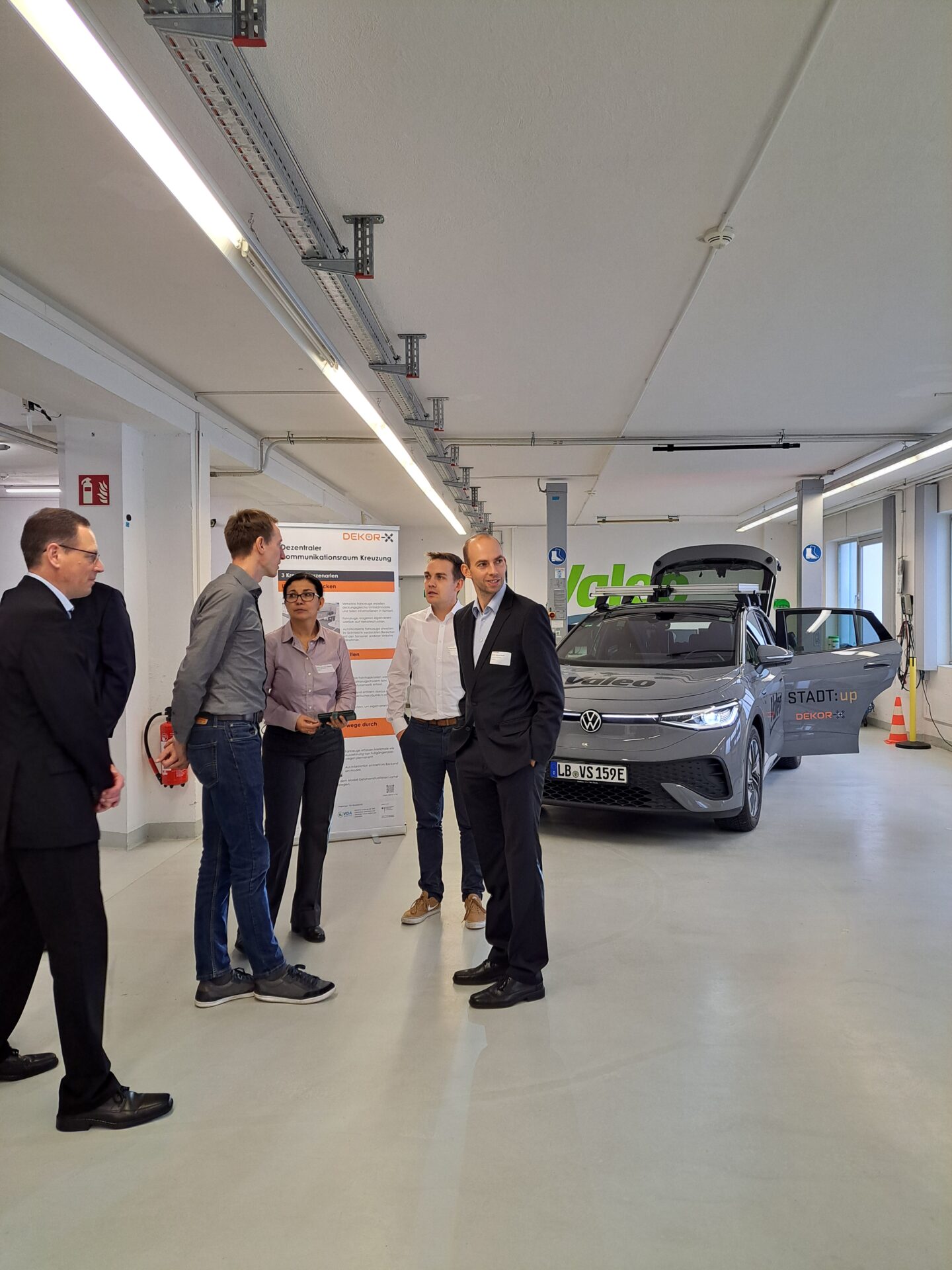 Mehrere Personen in Geschäftskleidung diskutieren über Smarte Kommunikation in einem hellen, geräumigen Raum, in dem ein Auto ausgestellt ist und ein großes DEKOR-X-Informationsplakat auf einer Staffelei über innovative Lösungen für sichere Straßen informiert. at Hochschule Coburg